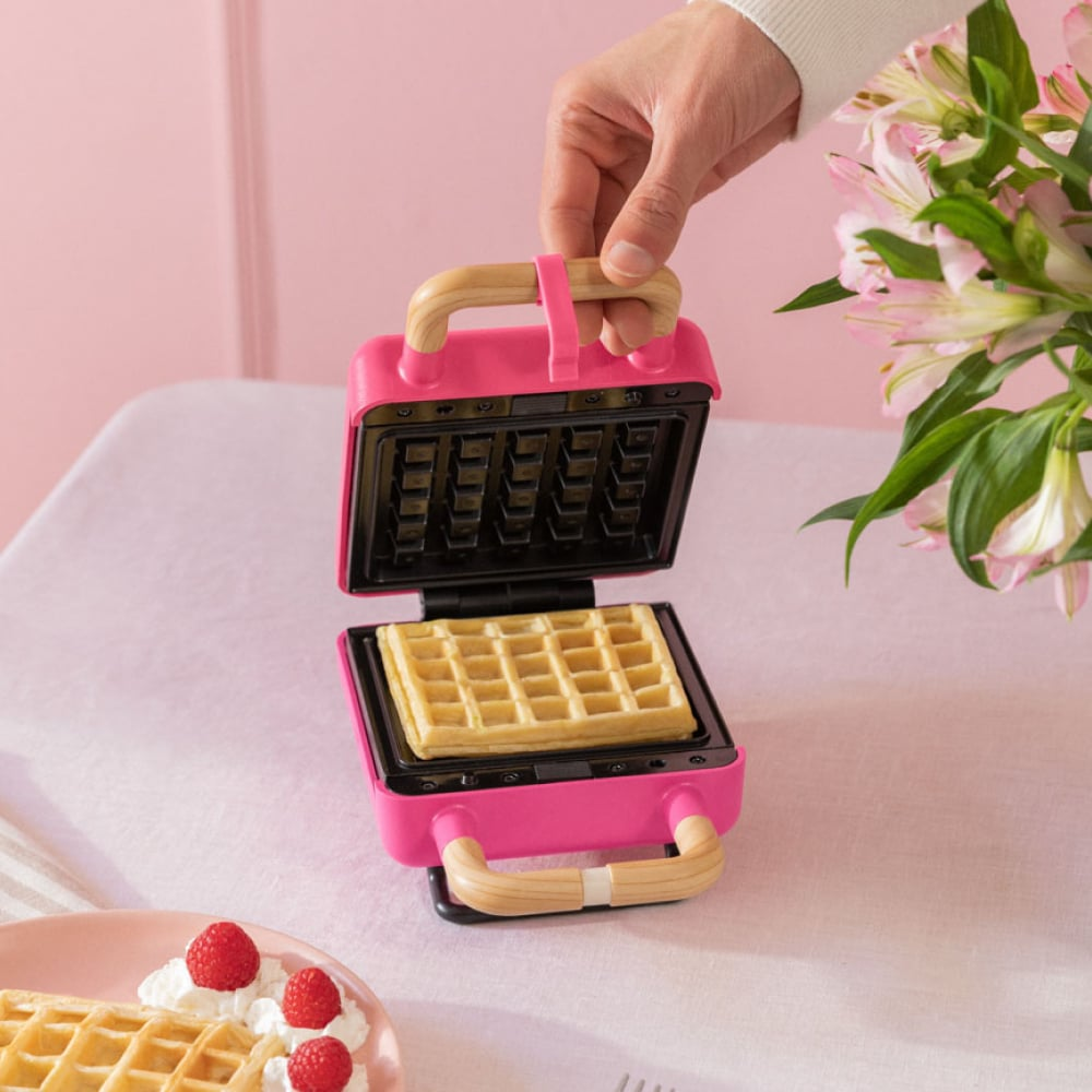 Máquina para hacer gofres rosa con un gofre dentro. Una mano lo va a cerrar. Más gofres en un plato.