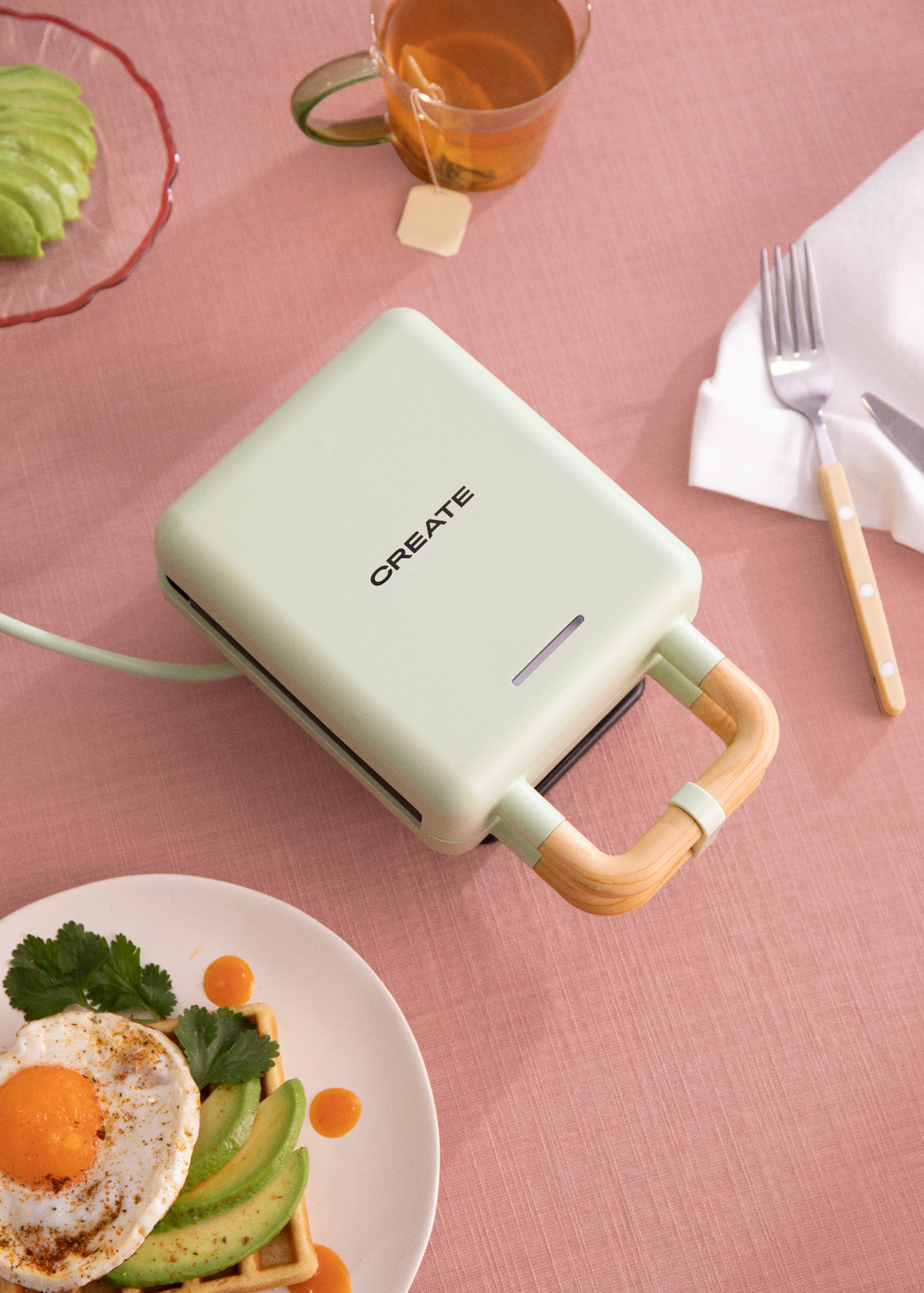 Una máquina para hacer gofres verde sobre una superficie rosa con alimentos de desayuno. Taza de té.
