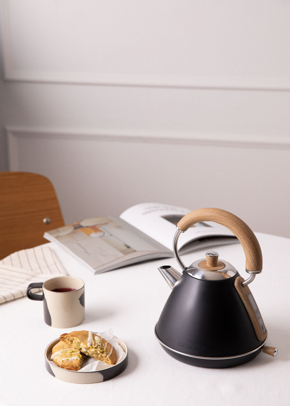 Una tetera negra con asa de madera sobre mesa con taza, pastel y revista.