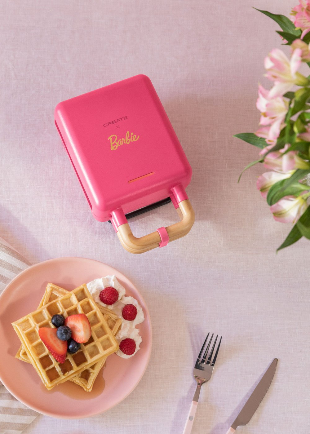 Una gofrera rosa Barbie con gofres, fruta y un tenedor. Un arreglo floral en el fondo.