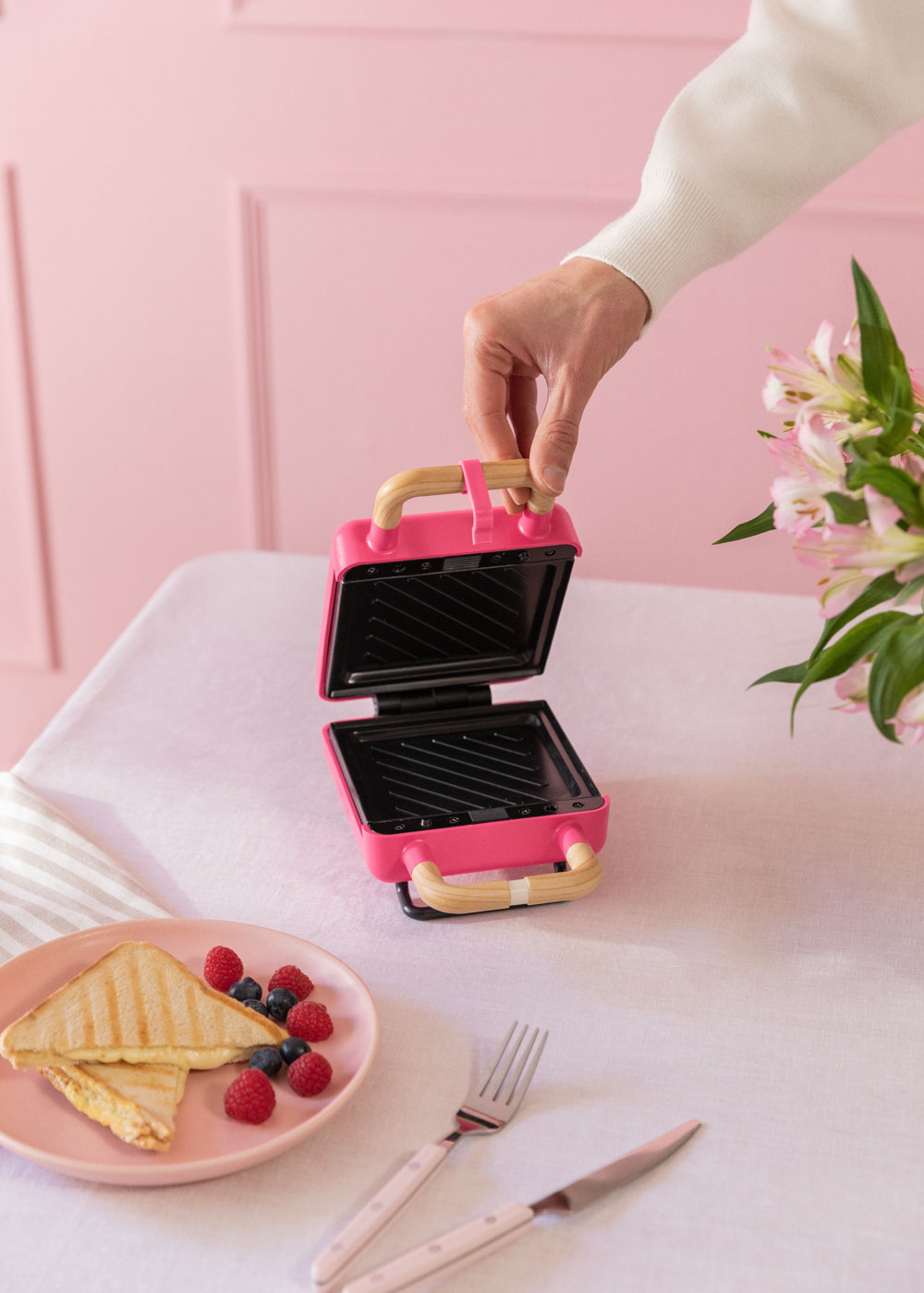 Máquina de gofres rosa sobre mesa blanca. Mano de persona alcanzando el mango.