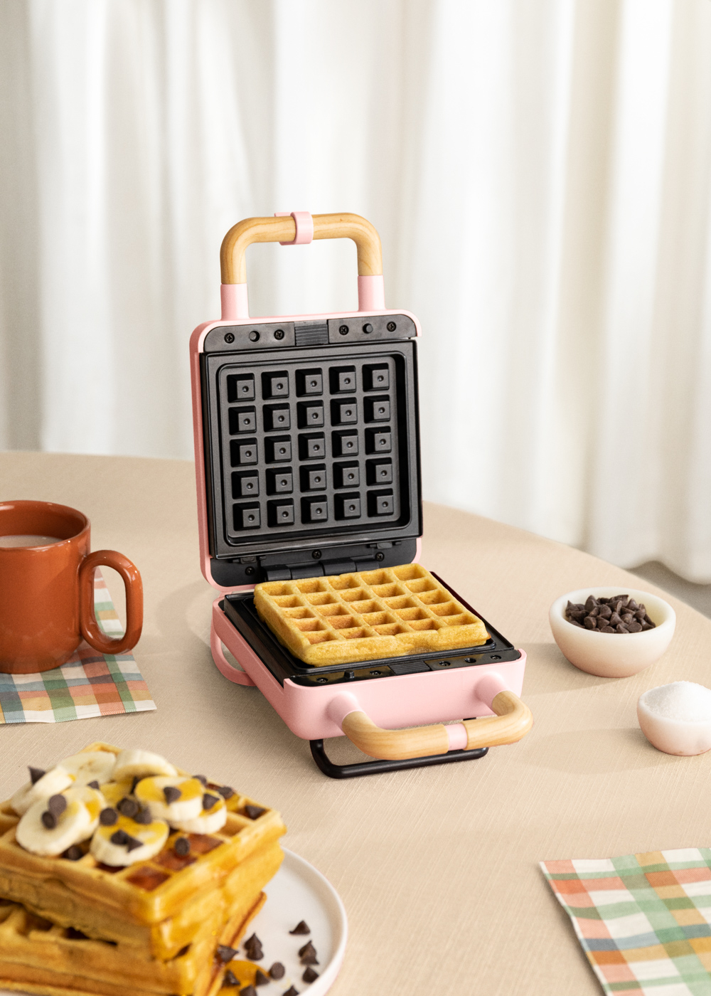 Una máquina de hacer gofres rosa con un gofre dentro. Gofres con plátano y chispas de chocolate cerca.