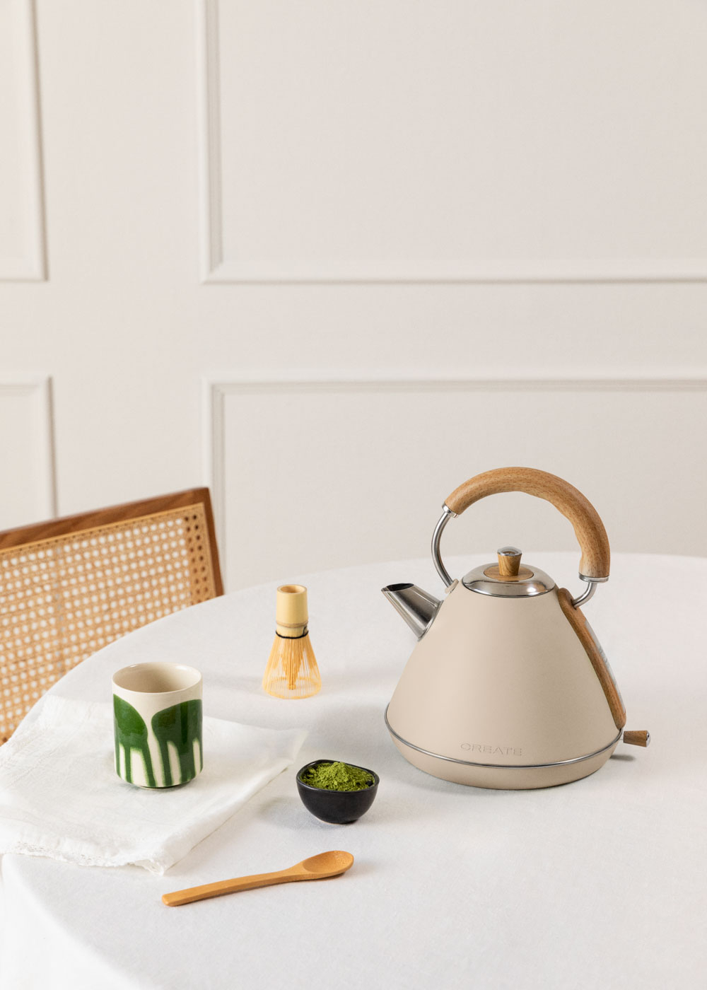Hervidor crema con asa de madera sobre mesa con accesorios de té. Fondo blanco.