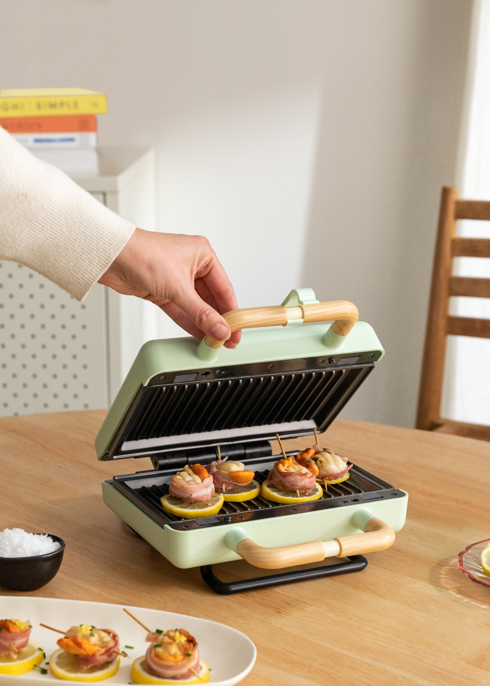 Una parrilla verde con comida dentro. Una mano la está cerrando. Brochetas con comida y sal en la mesa.