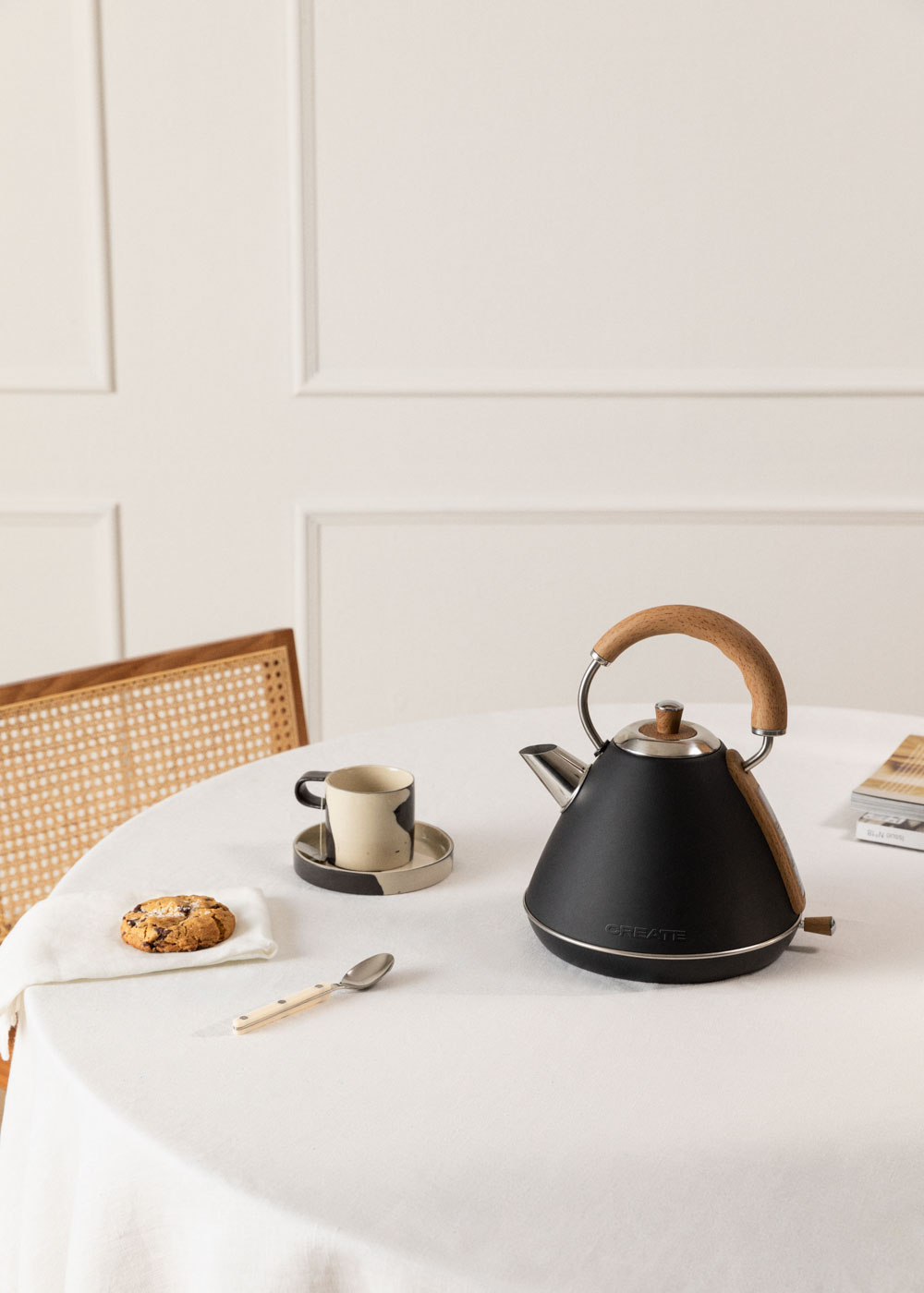 Schwarzer Wasserkocher auf weißer Tischdecke. Tasse und Keks daneben. Holzgriff.