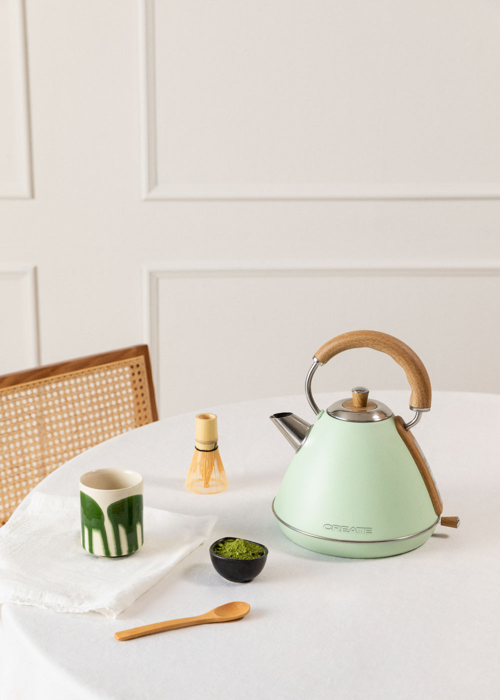 Küchenszene mit grünem Wasserkocher, Matcha-Set und einer Tasse auf weißer Tischdecke. Stuhl im Hintergrund.