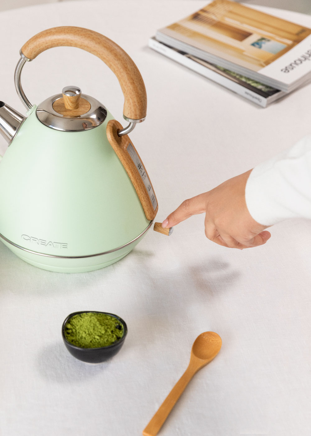 Eine Hand drückt einen Knopf an einem grünen Wasserkocher. Eine Schüssel mit Matcha und ein Löffel sind vorhanden.
