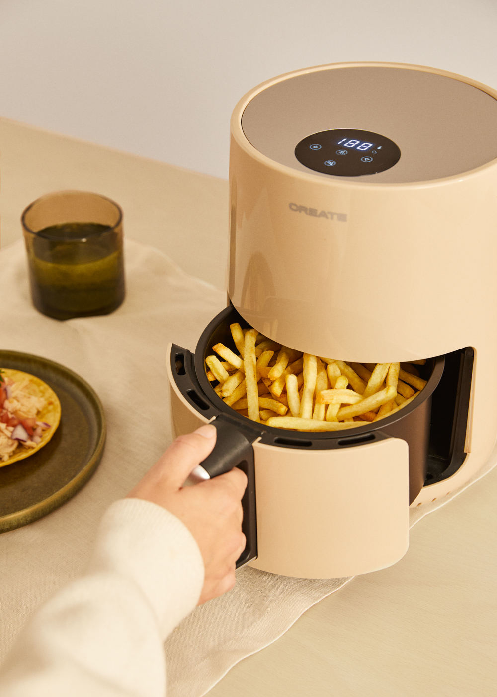 Freidora de aire beige con patatas fritas. Mano sacando la cesta. Vaso y taco a la izquierda.