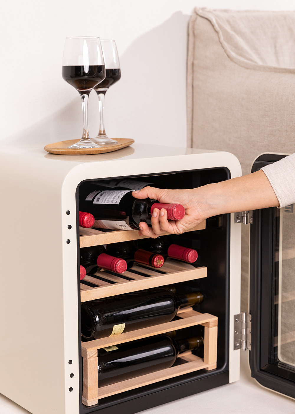 Una vinoteca color crema. Tiene botellas de vino dentro. Una mano alcanza una botella.