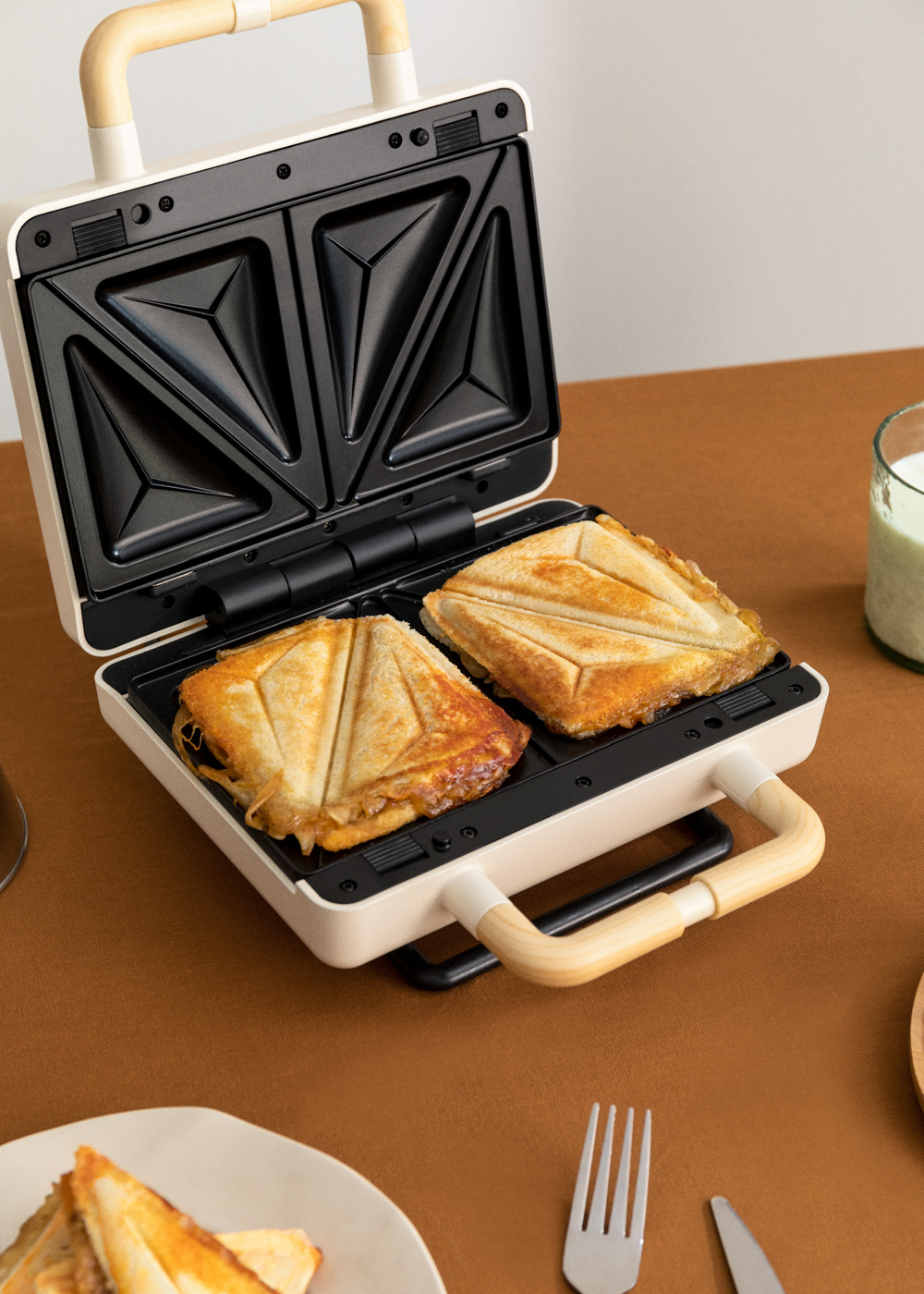 Sandwichera con 2 sándwiches tostados sobre superficie marrón, con bebida.
