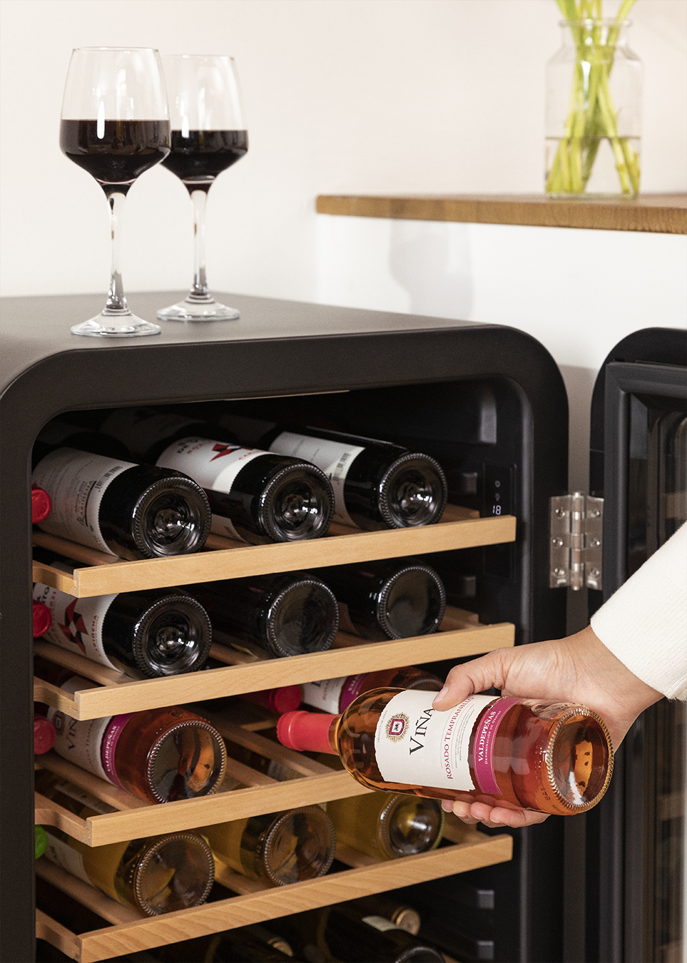 Una persona sacando una botella de vino de una vinoteca negra con copas al lado.