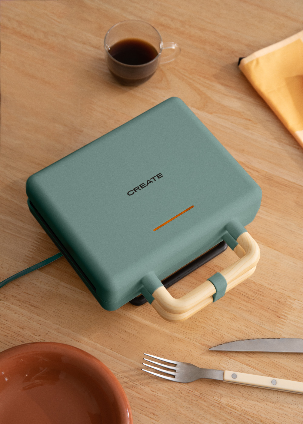 Máquina de gofres verde con mango de madera sobre mesa madera, café y plato.