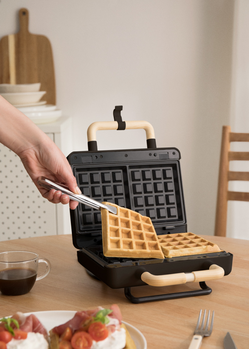 Mano usando pinzas para sacar un gofre cocido de una gofrera, con elementos de desayuno.