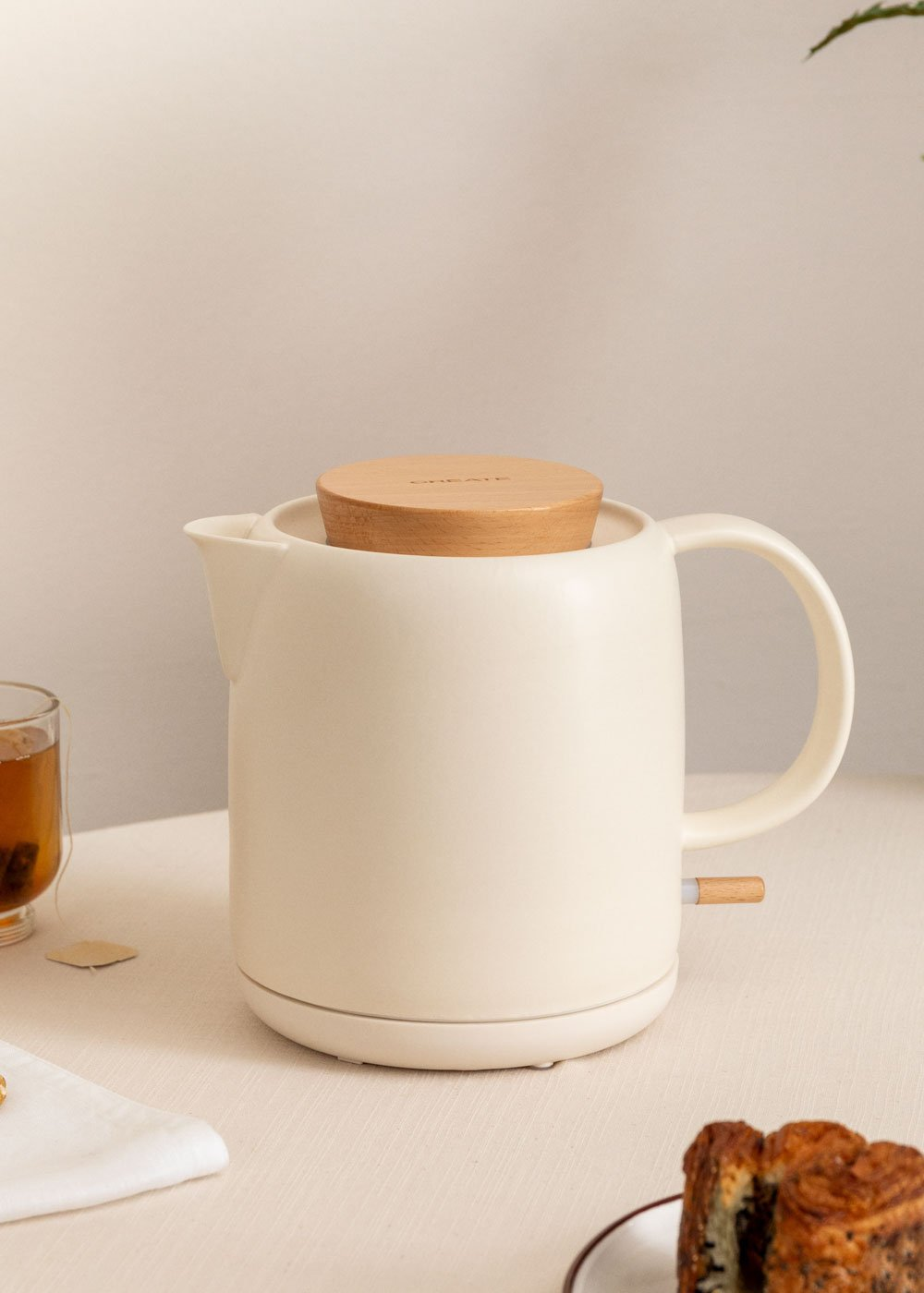 Hervidor eléctrico color marfil con tapa y asa de madera. Se ven una taza de té y pastelería.