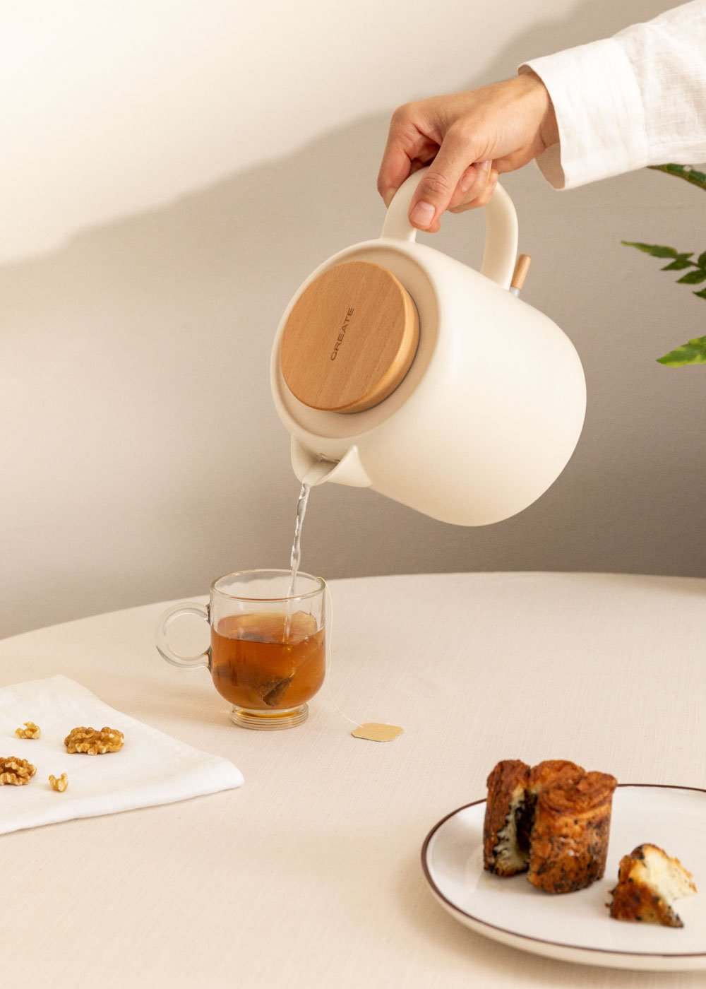 Una persona sirviendo té de una tetera blanca en un vaso. Hay nueces y pasteles en la mesa.