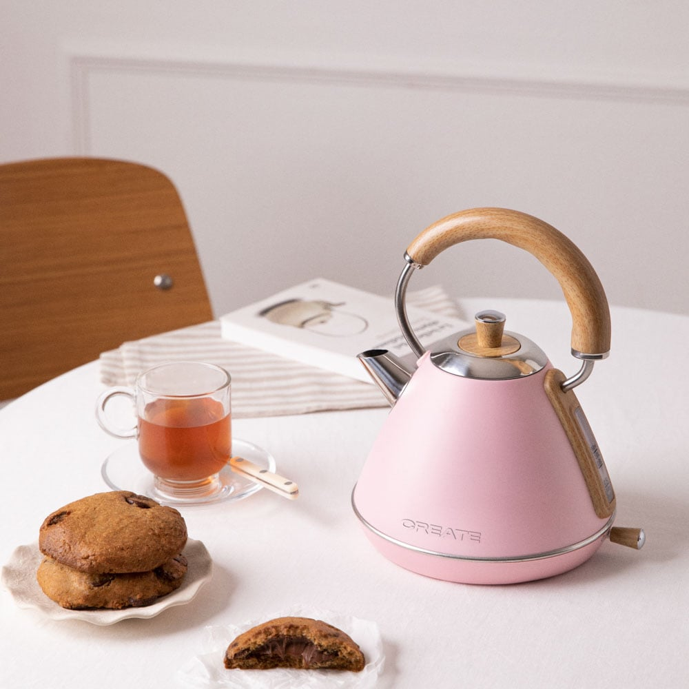 Rosa Wasserkocher mit Holzgriff auf weißem Tisch mit Keksen und Tee.