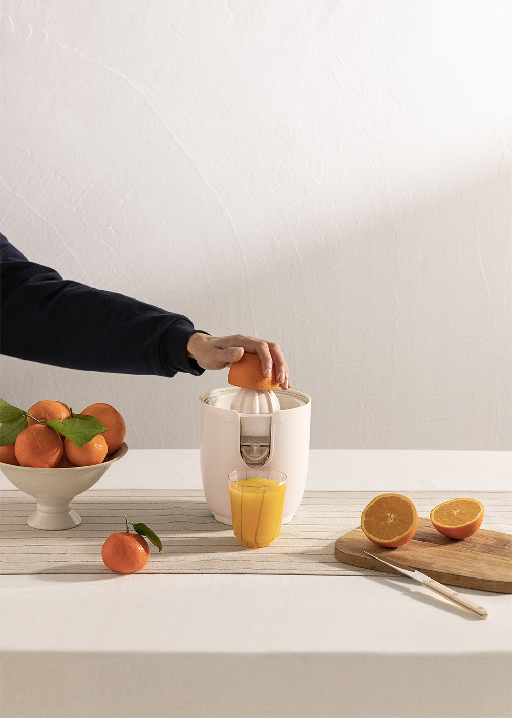 Persona exprimiendo una naranja. Exprimidor blanco. Naranjas en cuenco. Vaso de zumo de naranja.