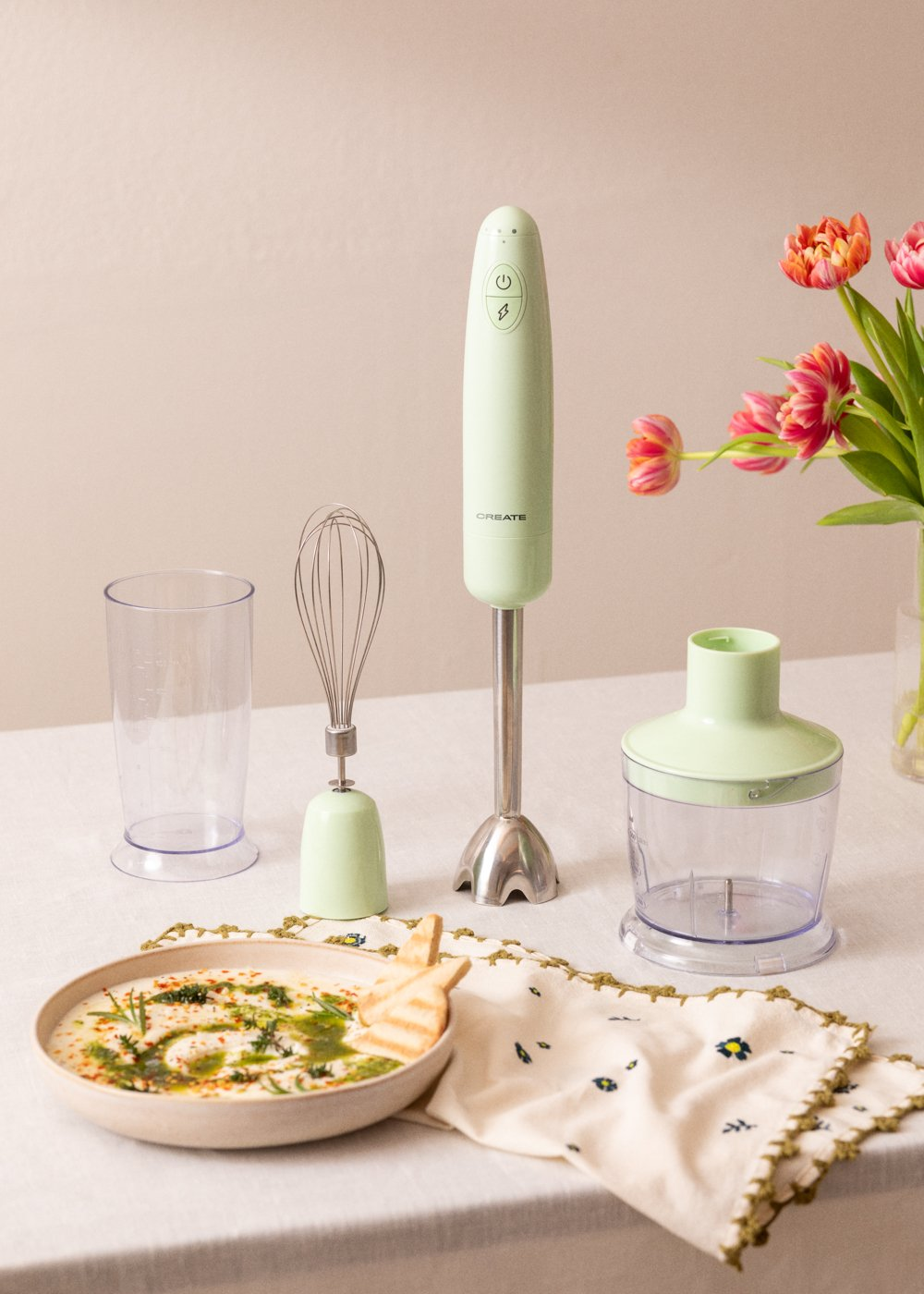 Una batidora de mano y accesorios sobre una mesa con un cuenco de comida y flores.