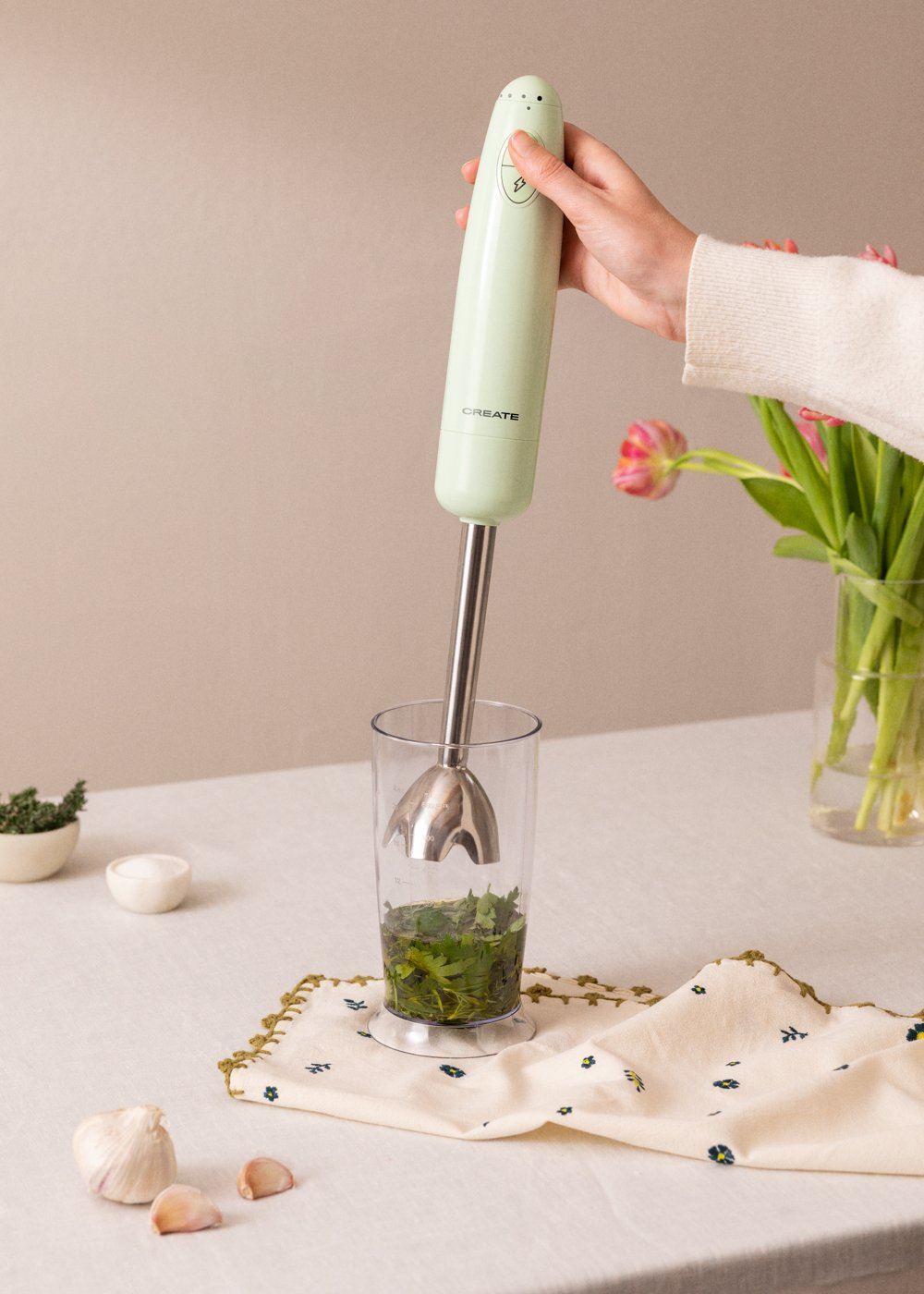 Persona usando una batidora de mano verde. Vaso con hierbas, ajo y flores al fondo.