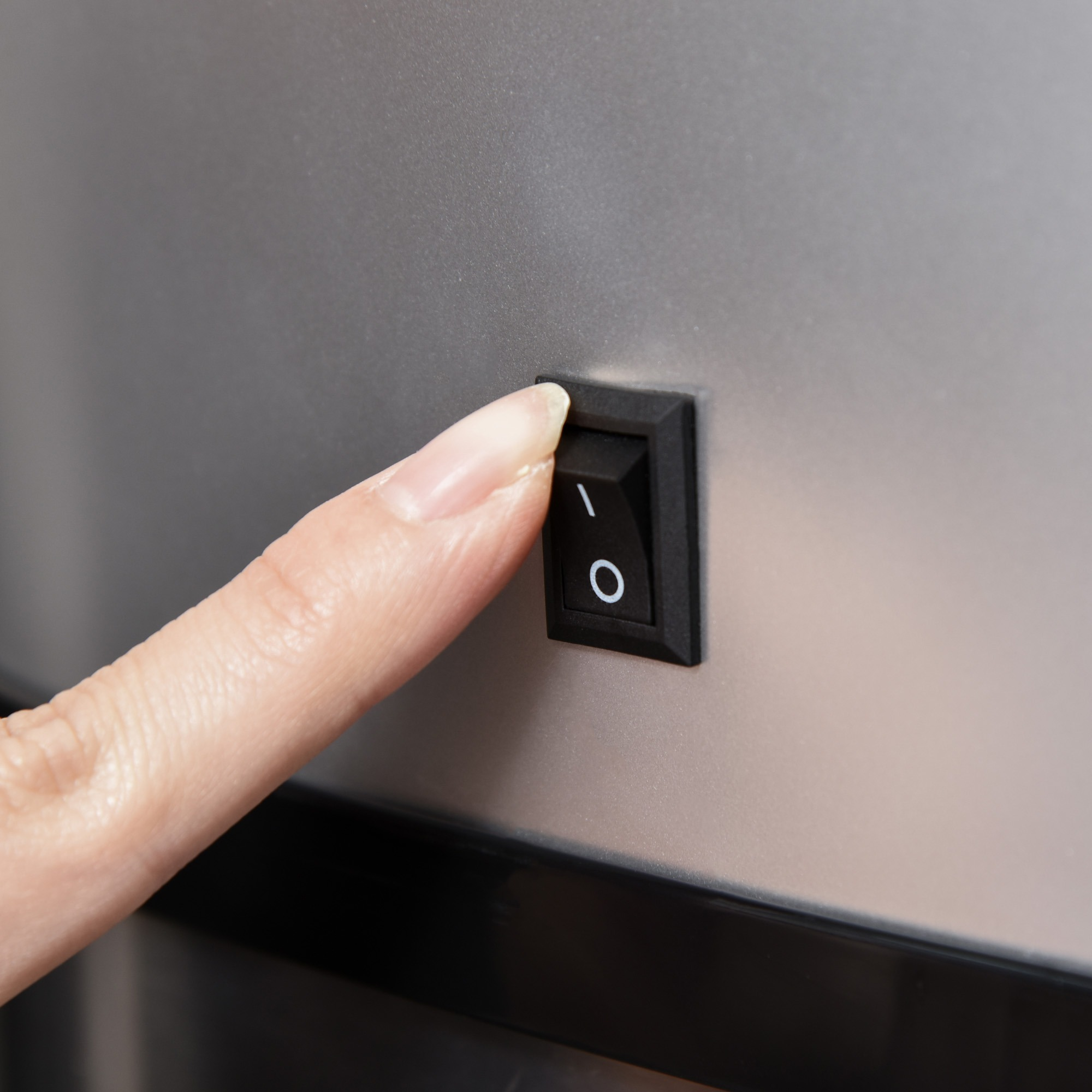 Ein Finger drückt einen schwarzen Ein-/Ausschalter mit „I/O“-Symbol auf grauer Oberfläche.