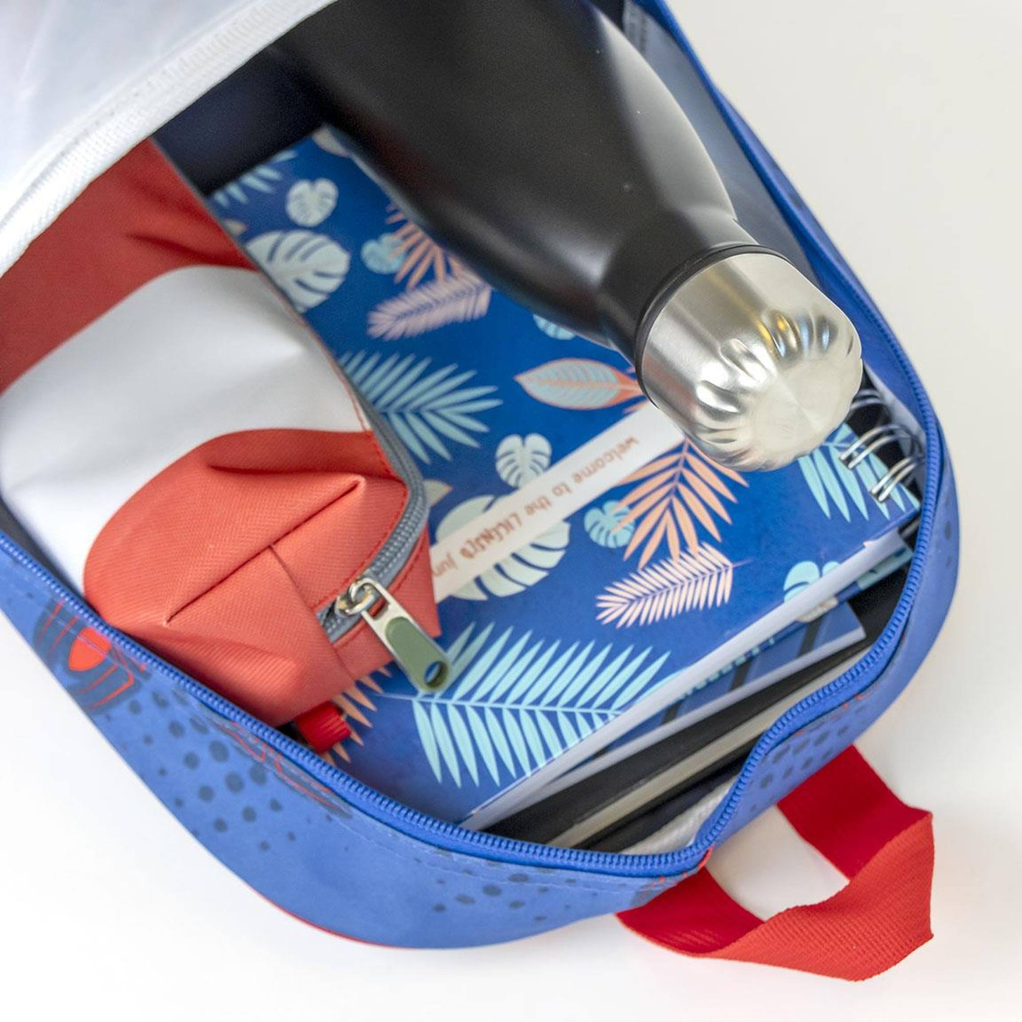 Blauer Rucksack geöffnet, gefüllt mit einer schwarzen Wasserflasche, Notizbuch und Federmäppchen. Weißer Hintergrund.