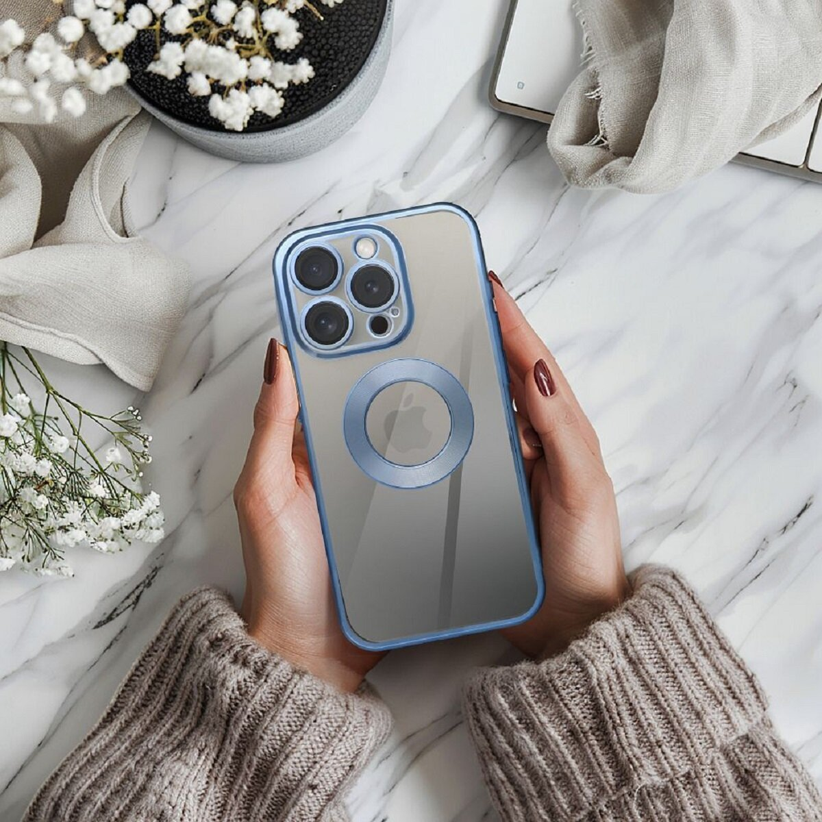 Hände halten ein Telefon mit einer blauen Hülle. Das Telefon liegt auf einer Marmoroberfläche, eine Vase und eine Tastatur sind im Hintergrund.