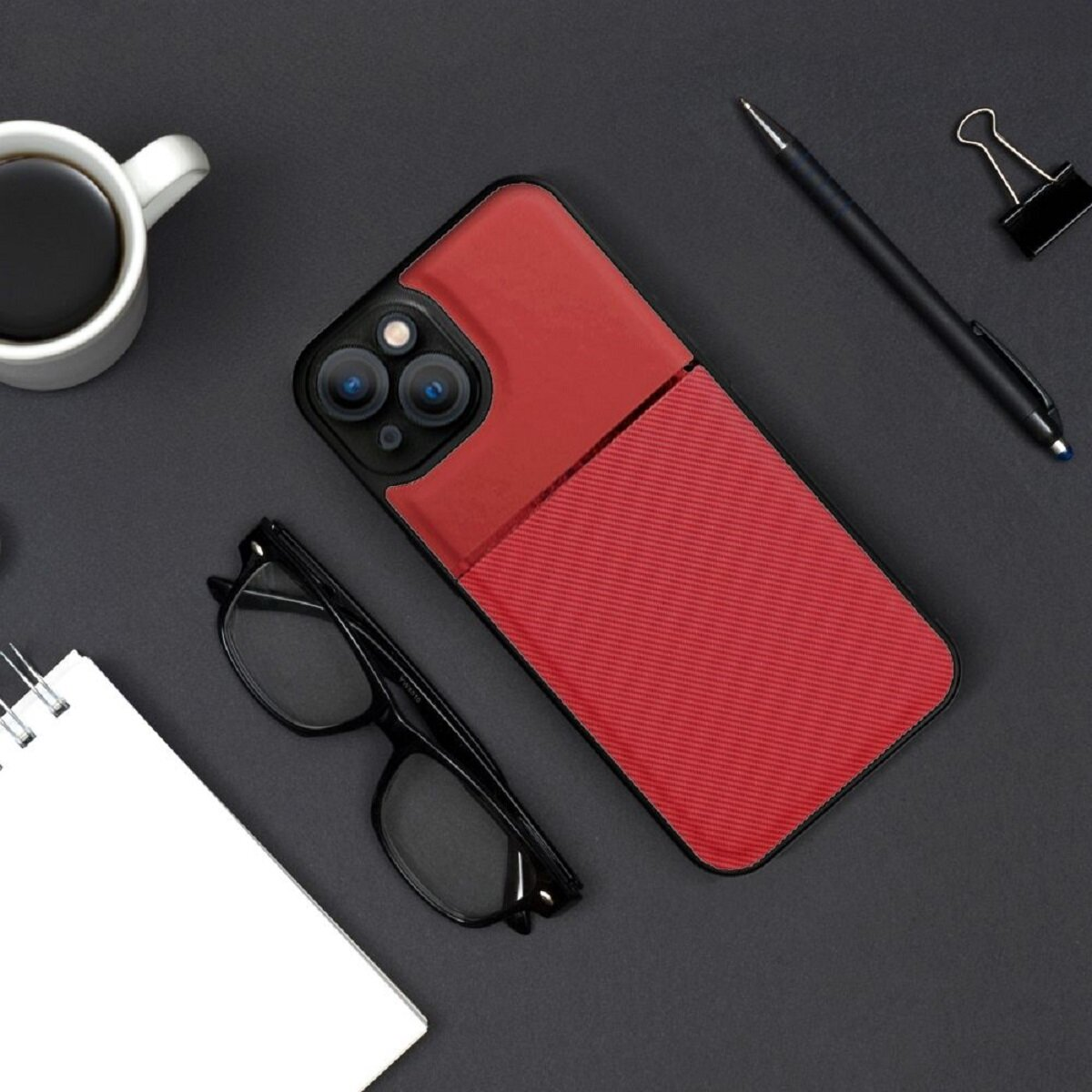 Schreibtisch-Setup mit roter Handyhülle, Brille, Notizbuch, Becher, Stift und Büroklammer.