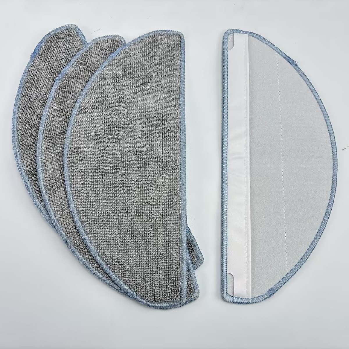 Cztery szare nakładki do mopa z mikrofibry i jedna biała z niebieskim obszyciem, na białej powierzchni.