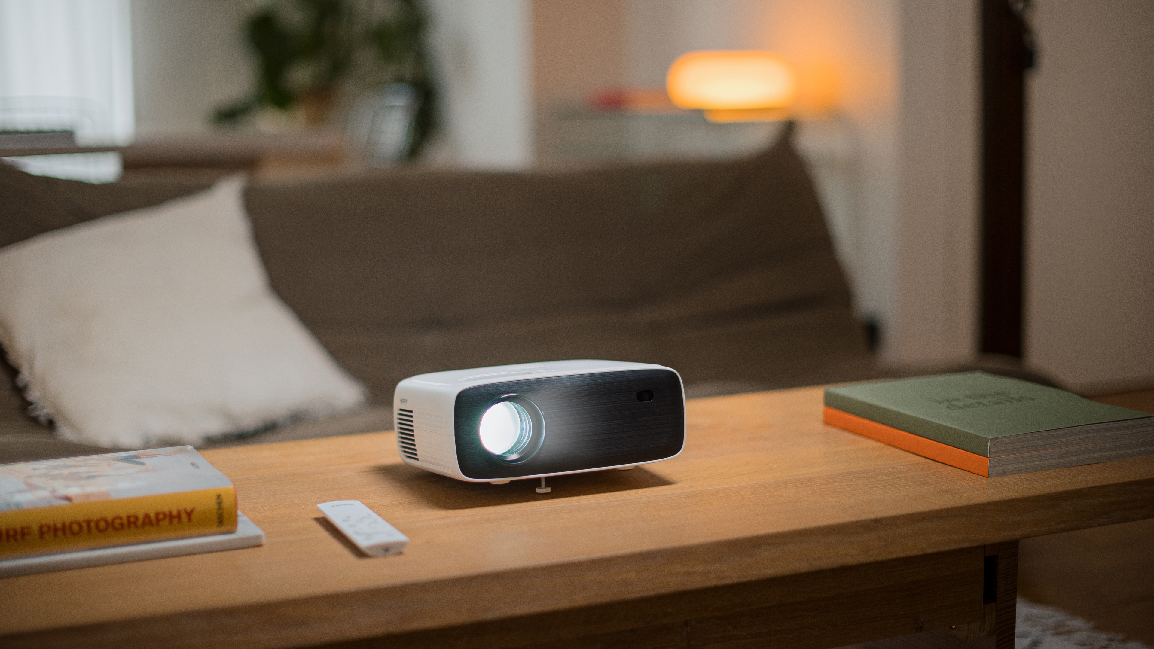 Projektor na drewnianym stole. Książki i pilot w pobliżu. Sofa i lampa w tle.