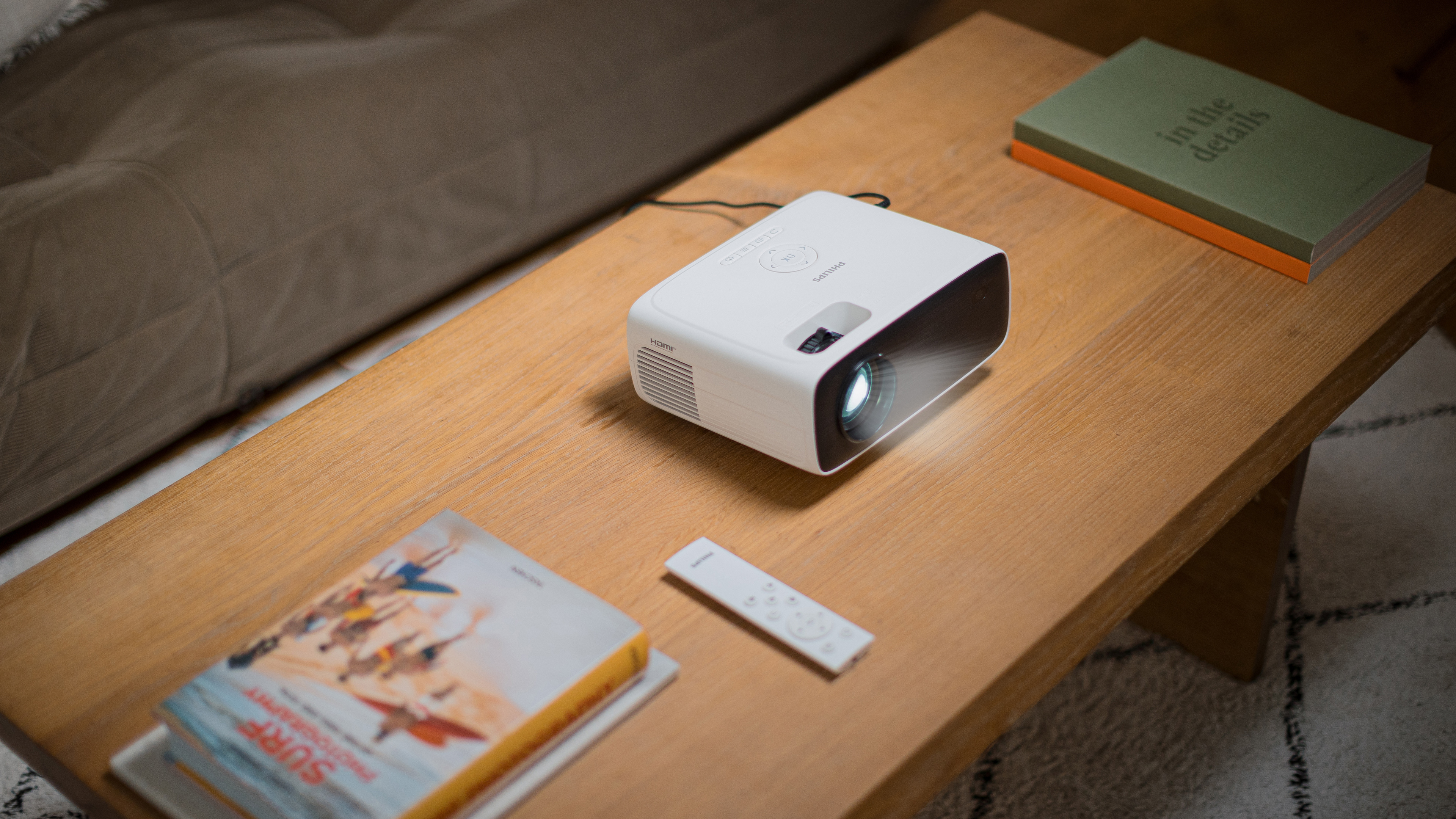 Biały projektor na drewnianym stole z książkami i pilotem. Widoczne światło projekcji.