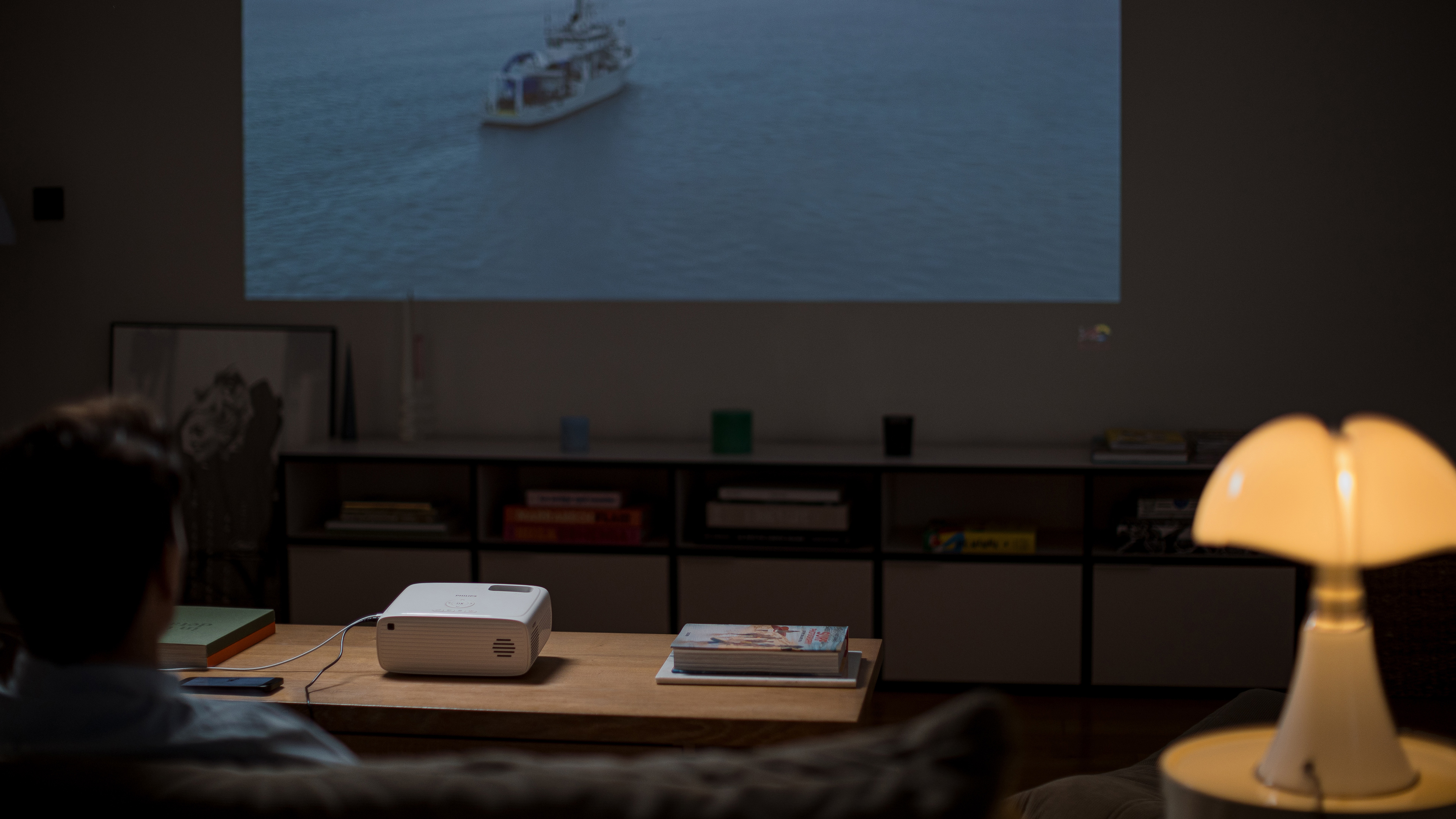 Osoba oglądająca film wyświetlany na ścianie. Lampa i stół na pierwszym planie.