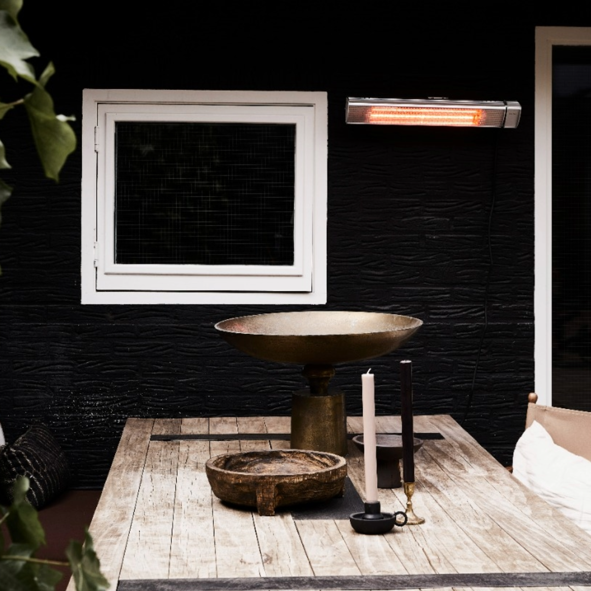 Scena all'aperto con tavolo in legno, candele e riscaldatore. Muro nero con finestra bianca. Riscaldatore sul muro.
