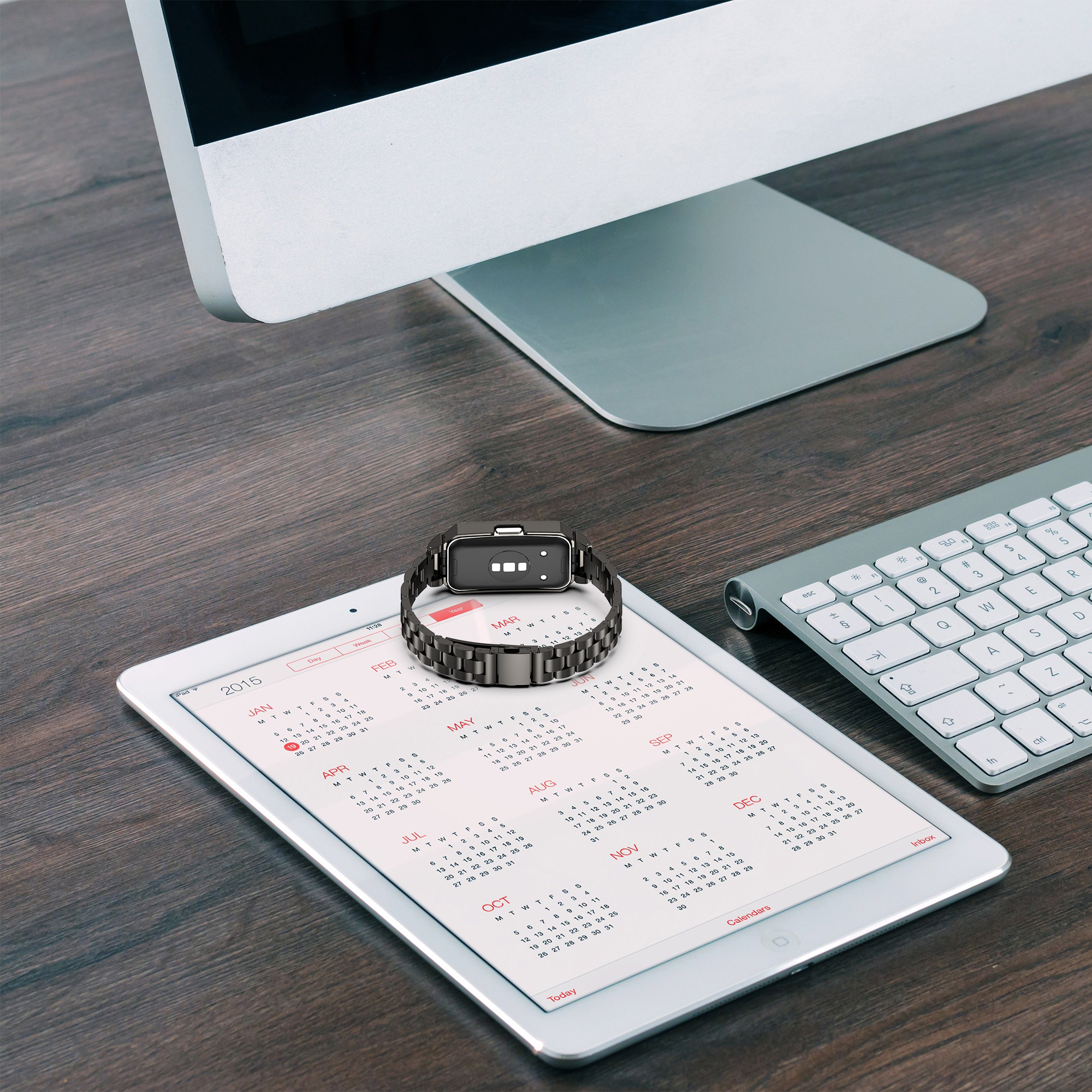 Eine schwarze Smartwatch auf einem Tablet, das einen Kalender anzeigt. Tastatur und Monitor sind zu sehen.