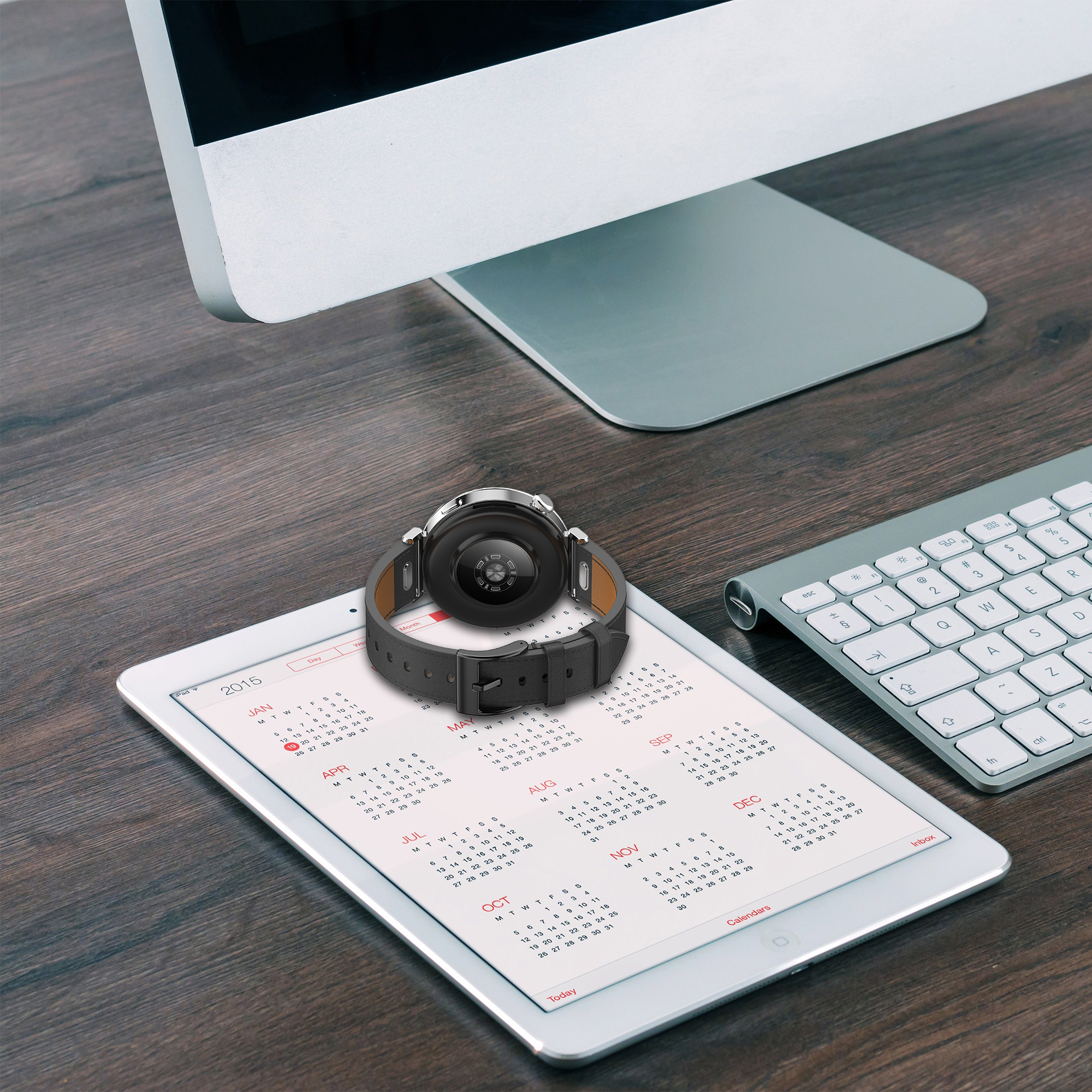 Uno smartwatch con cinturino in pelle su un tablet con calendario, e una tastiera, con un monitor sullo sfondo.