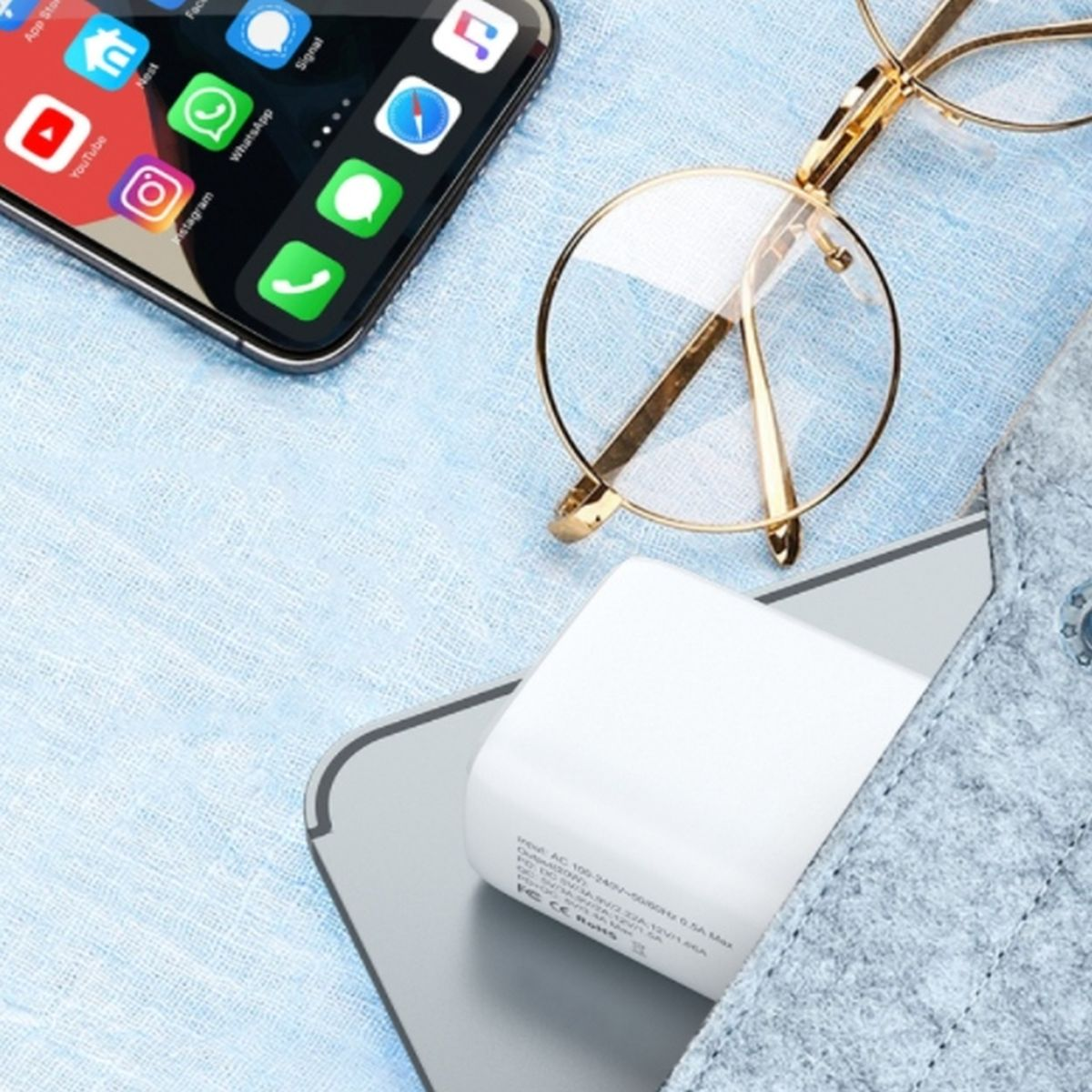 Telefon z aplikacjami, okularami i białą ładowarką na jasnoniebieskiej tkaninie. Szare etui jest obecne.