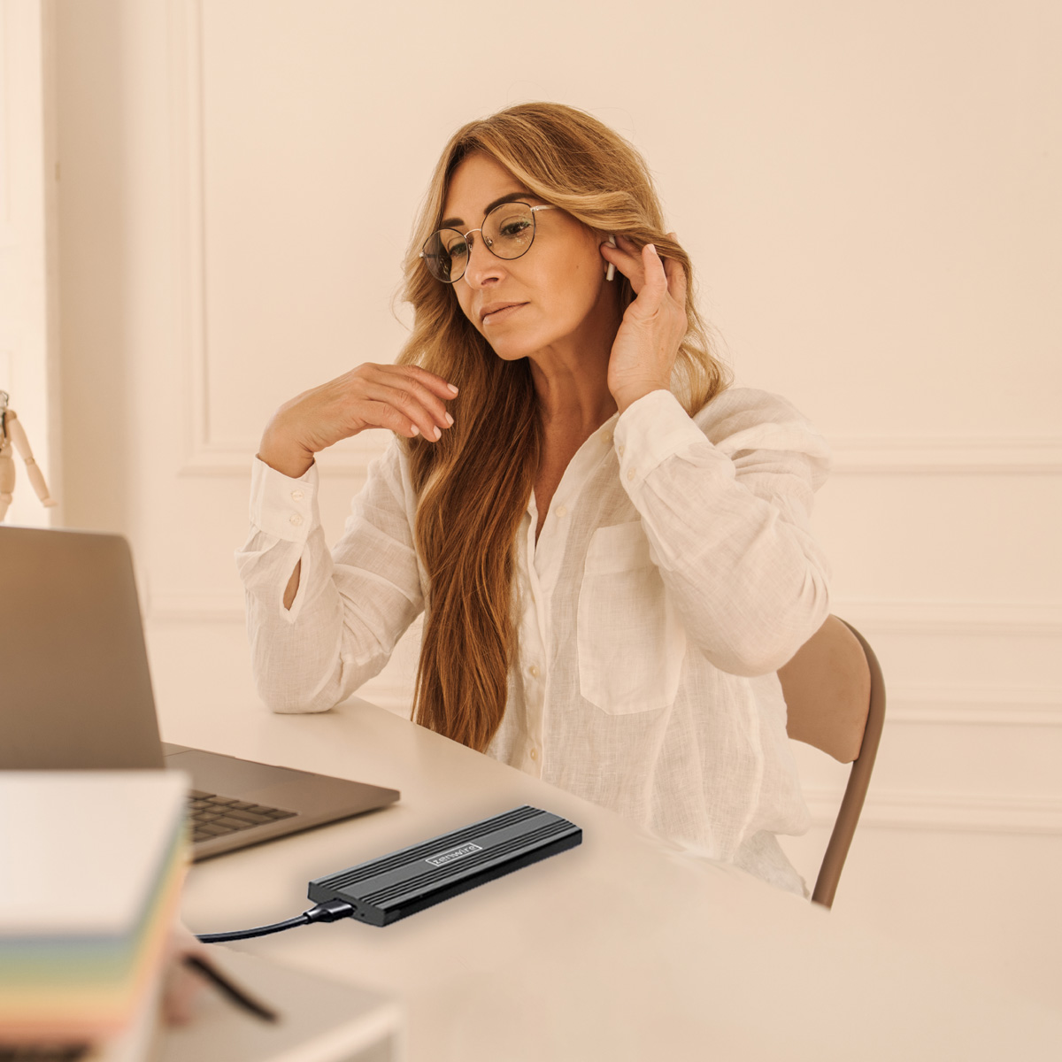 Kobieta w okularach przy biurku. Jest laptop i czarne urządzenie. Reguluje słuchawkę.