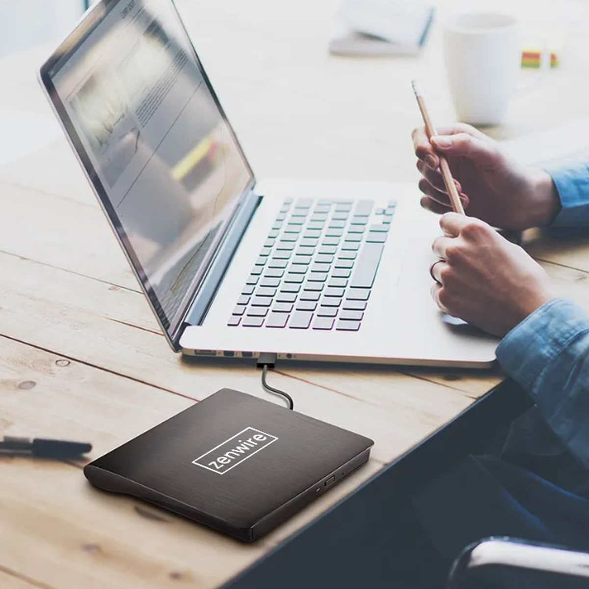 Czarny dysk zewnętrzny Zenwire jest podłączony do laptopa na drewnianym biurku. Osoba trzyma ołówek.