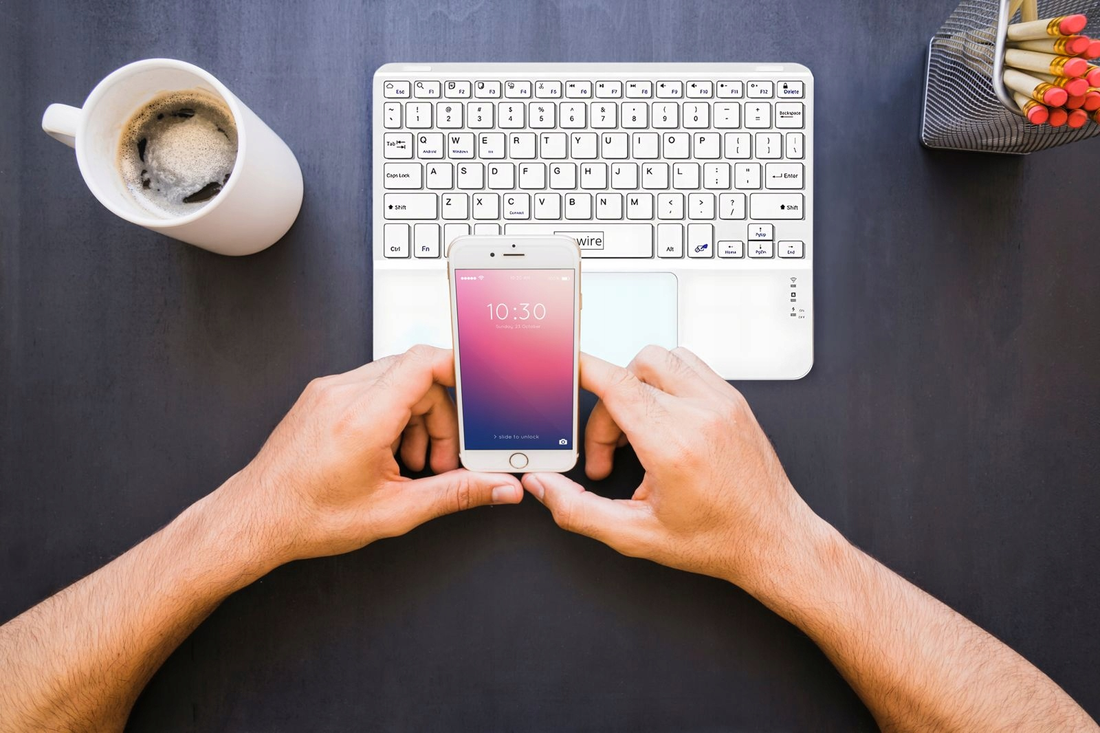 Dłonie trzymają telefon na białej klawiaturze, obok filiżanki kawy i ołówków.