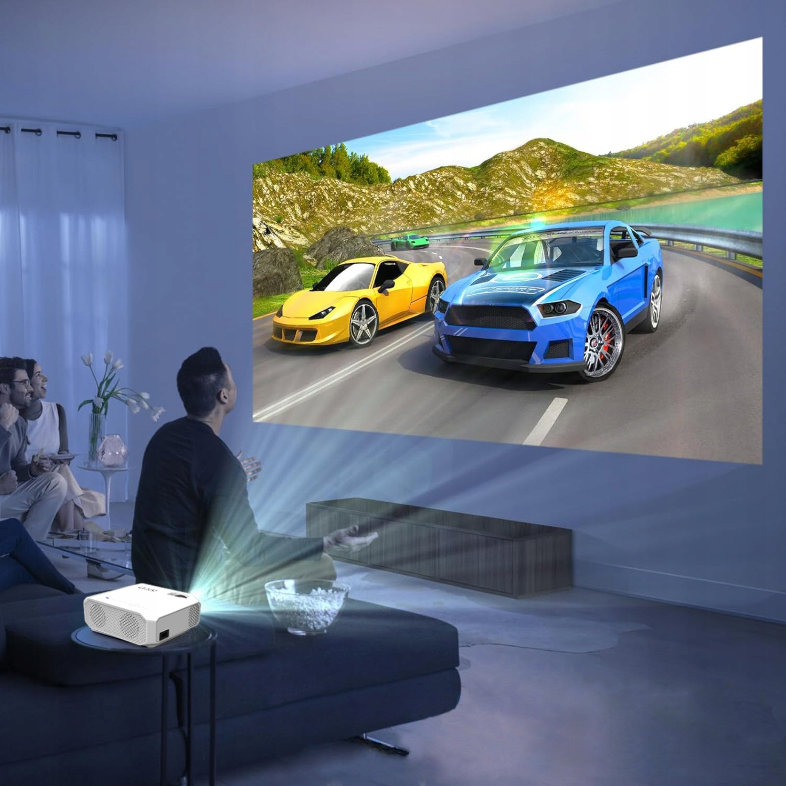 Projektor wyświetla wyścig samochodowy na dużym ekranie. Ludzie oglądają. Projektor i miska na stole.