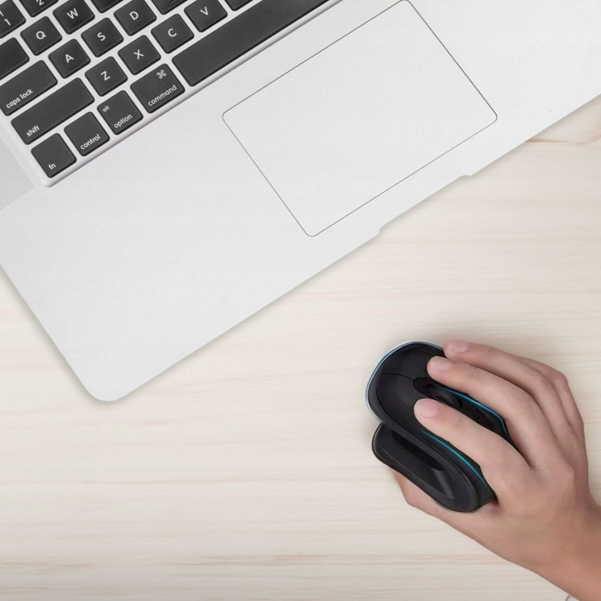 Ręka trzymająca czarną mysz obok laptopa na jasnym drewnianym stole.