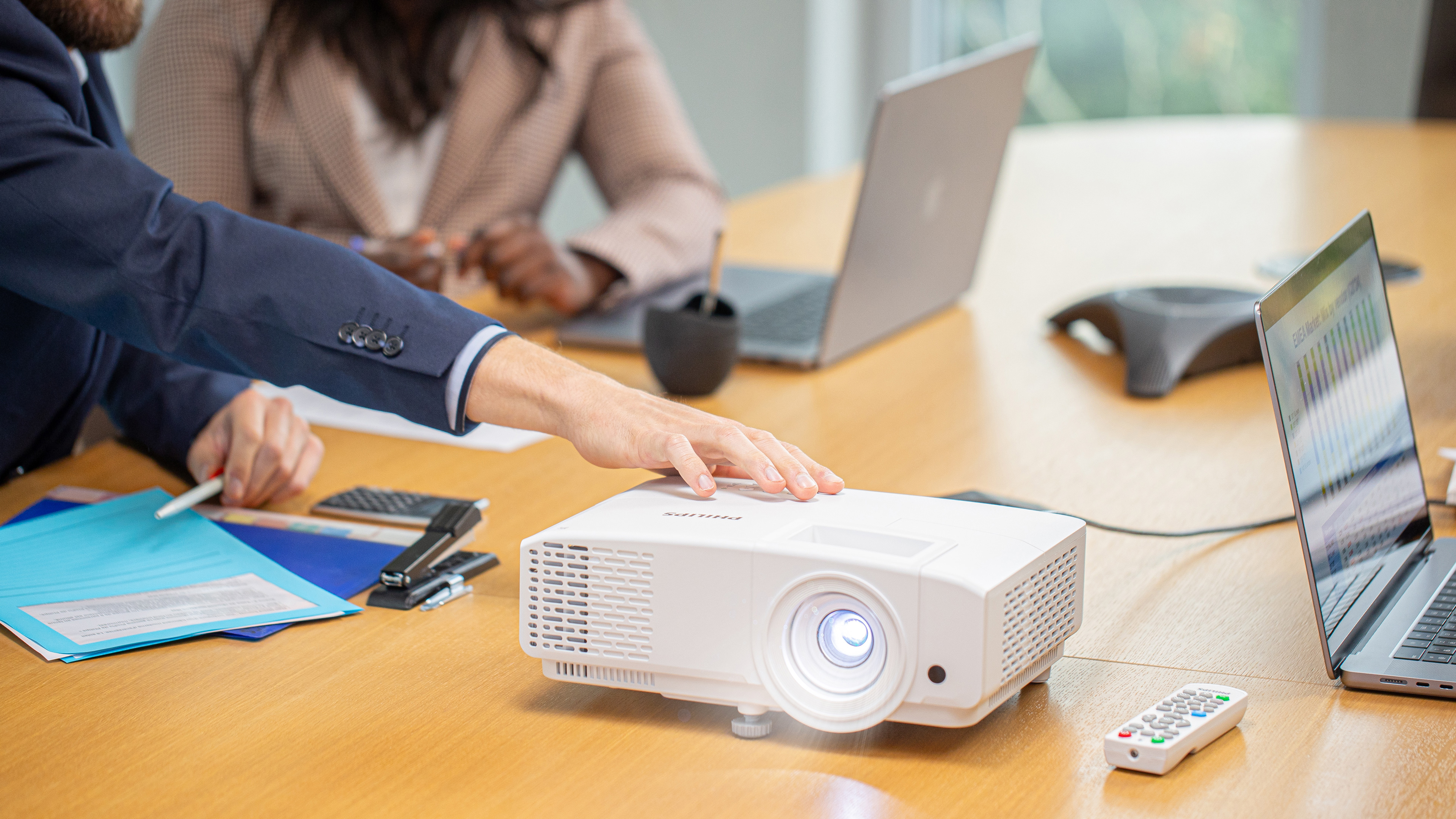 Twee mensen in een vergadering. Eén wijst naar een witte projector op een tafel. Laptops en een afstandsbediening zijn ook aanwezig.