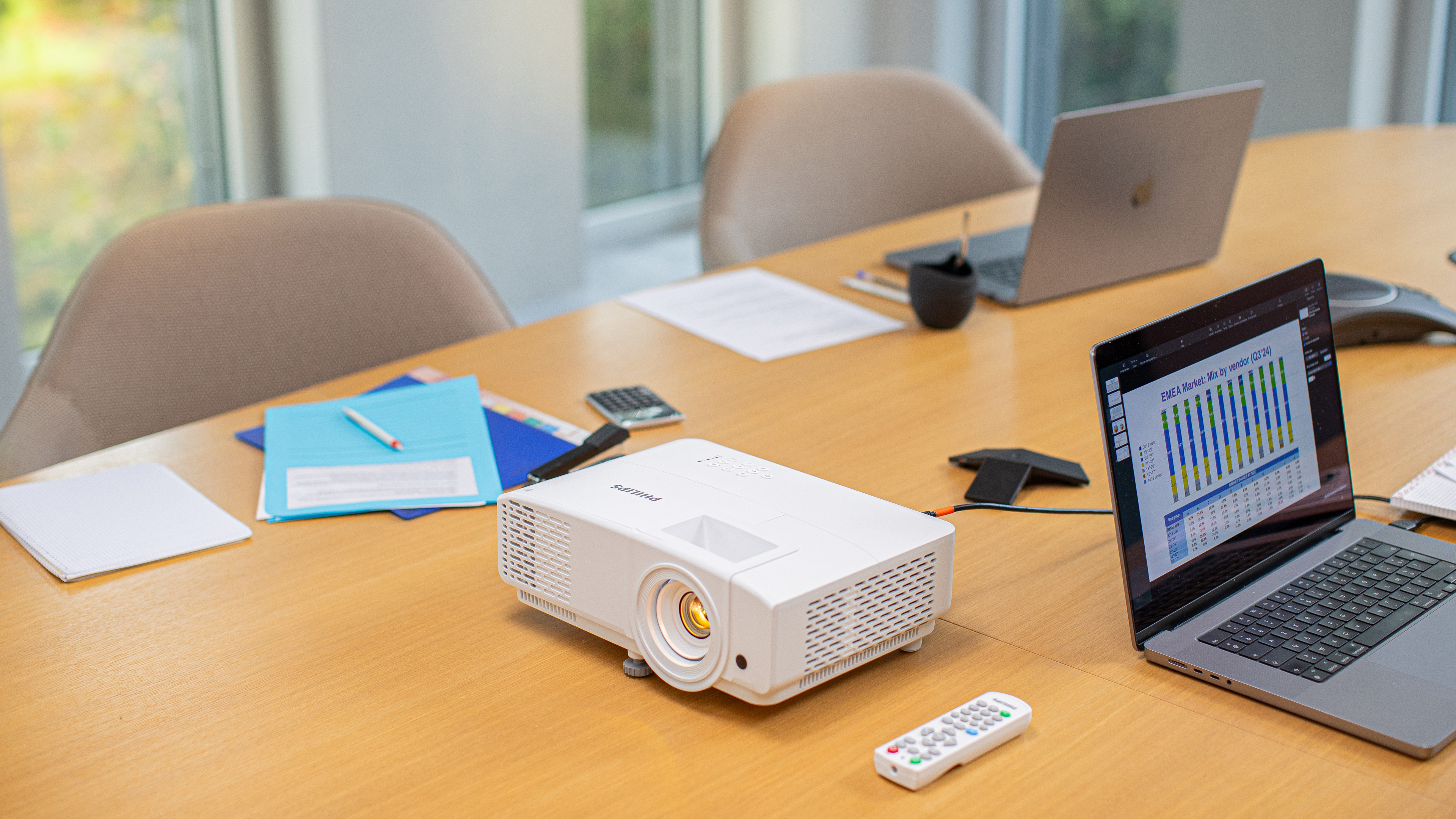 Een projector op een tafel. Laptops, papieren en een afstandsbediening liggen ook op de tafel.