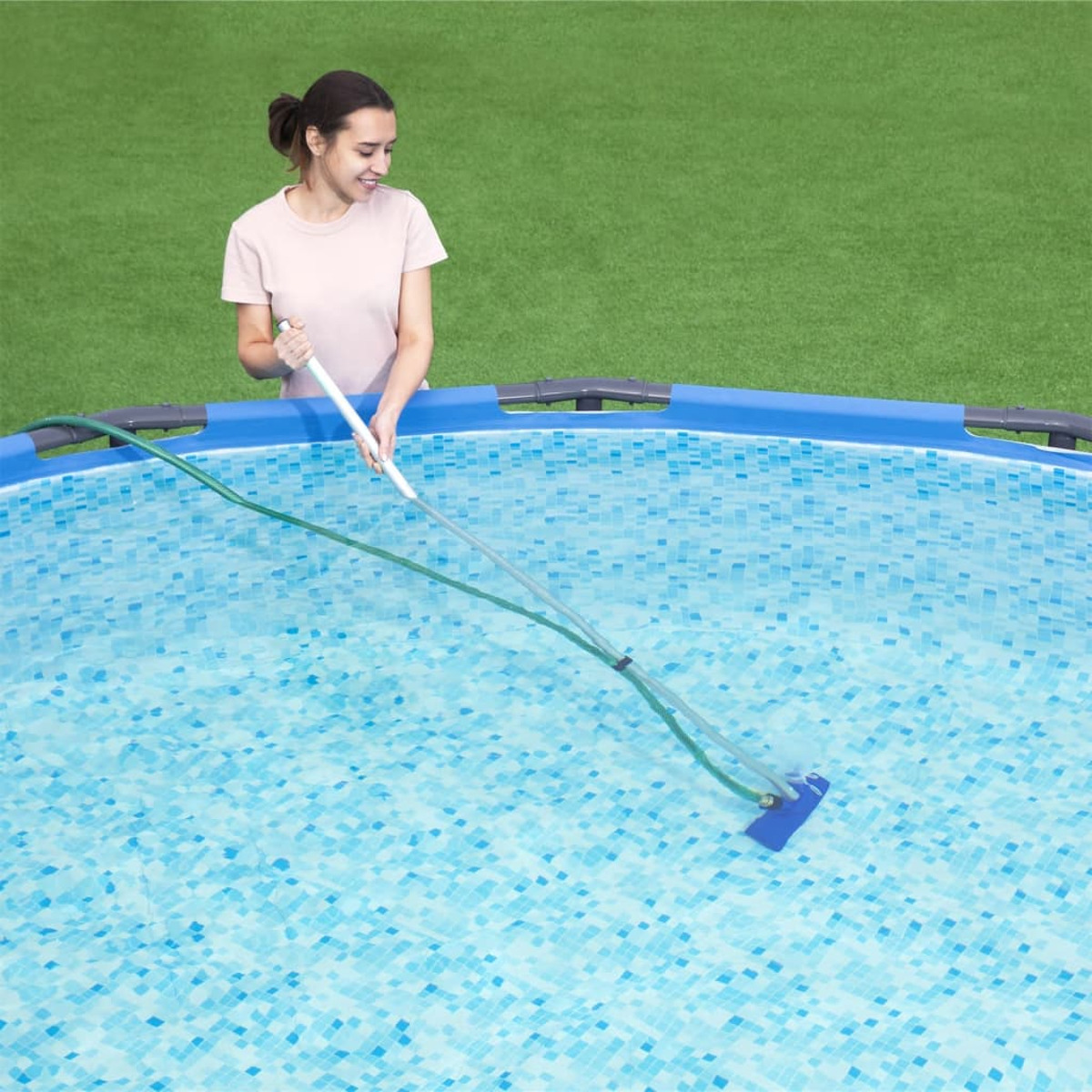Una donna pulisce una piscina con piastrelle blu con una spazzola a manico lungo. Erba verde sullo sfondo.