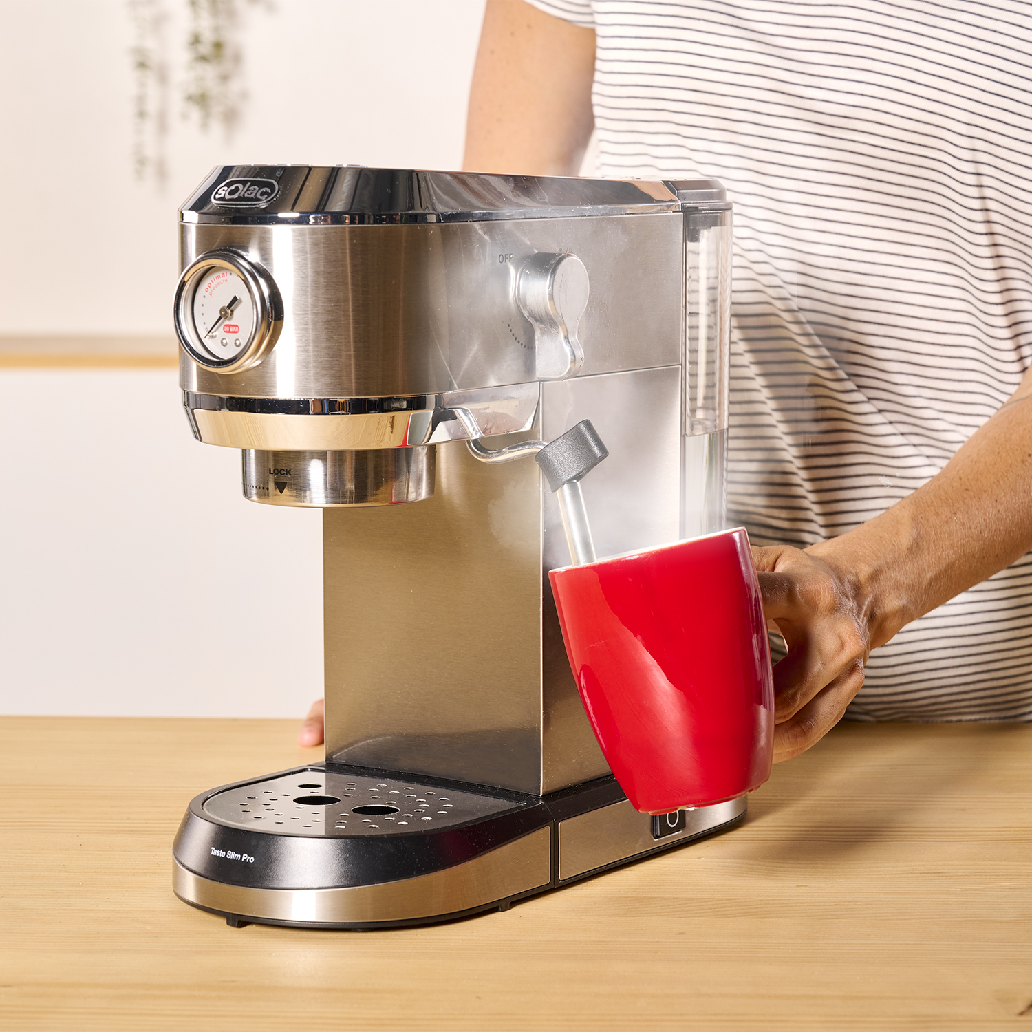 Una persona sosteniendo una taza roja debajo de una máquina de espresso plateada. Una mesa de madera.