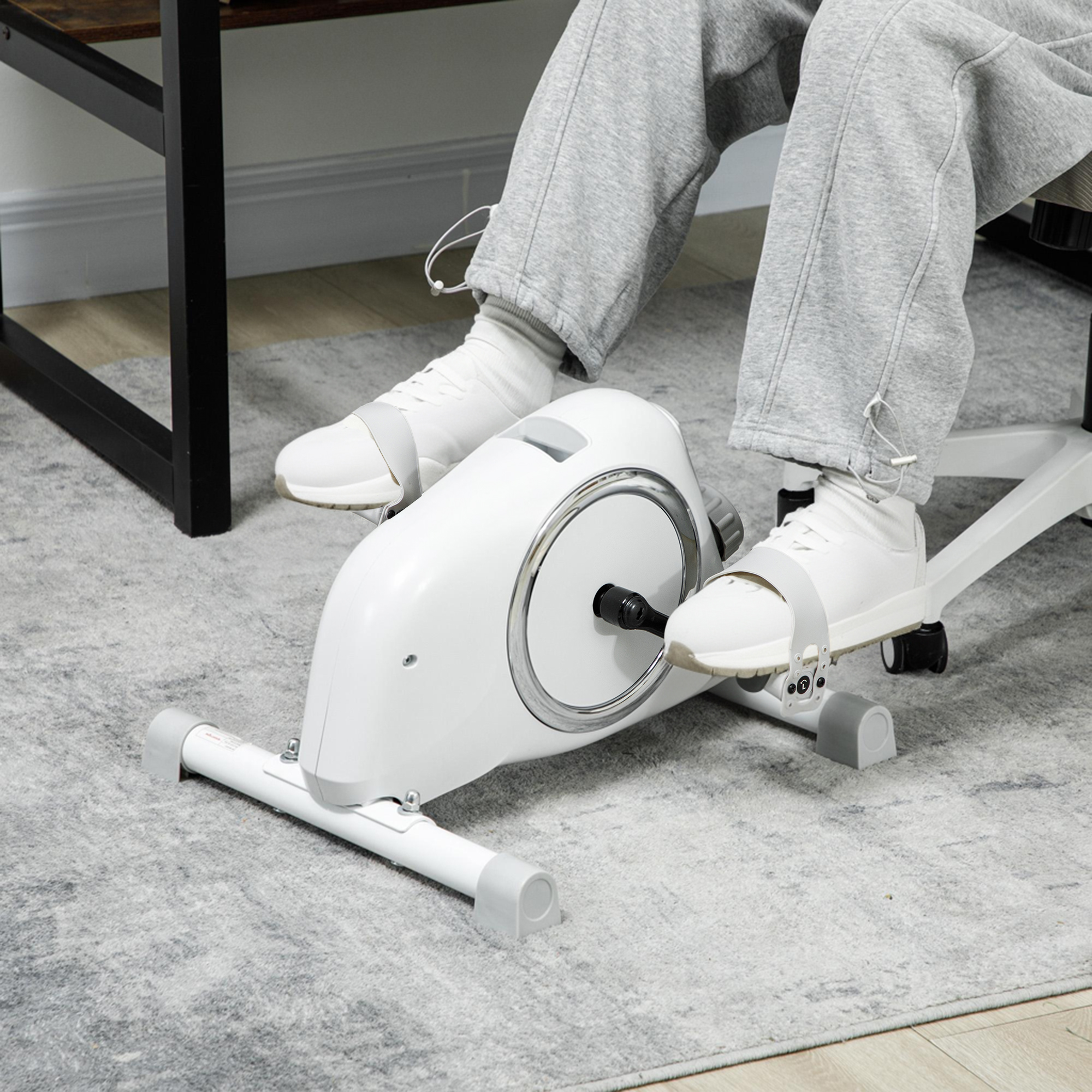 Eine Person benutzt ein Unter-Schreibtisch-Heimtrainer auf einem hellen Teppich. Weiße Turnschuhe sind sichtbar.