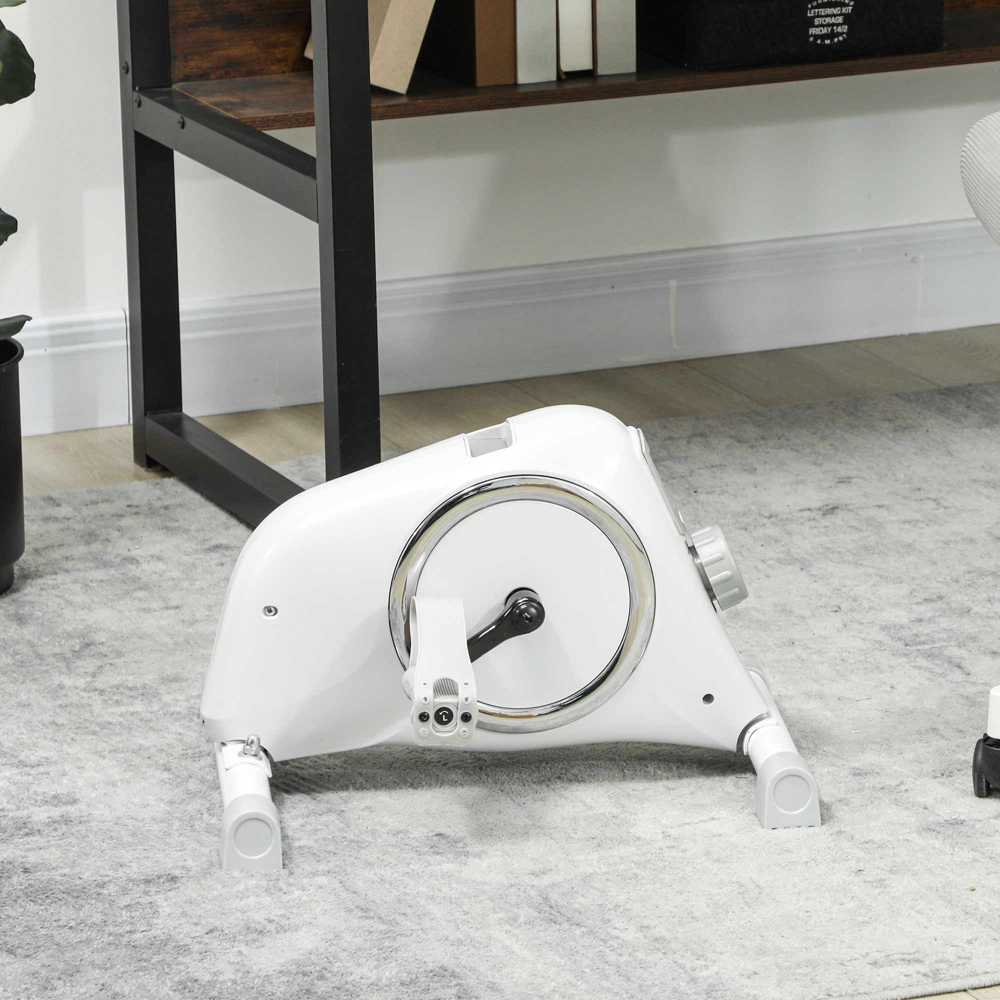 Ein weißes Mini-Ergometer auf einem Teppich. Es hat ein silbernes Rad und Pedale.