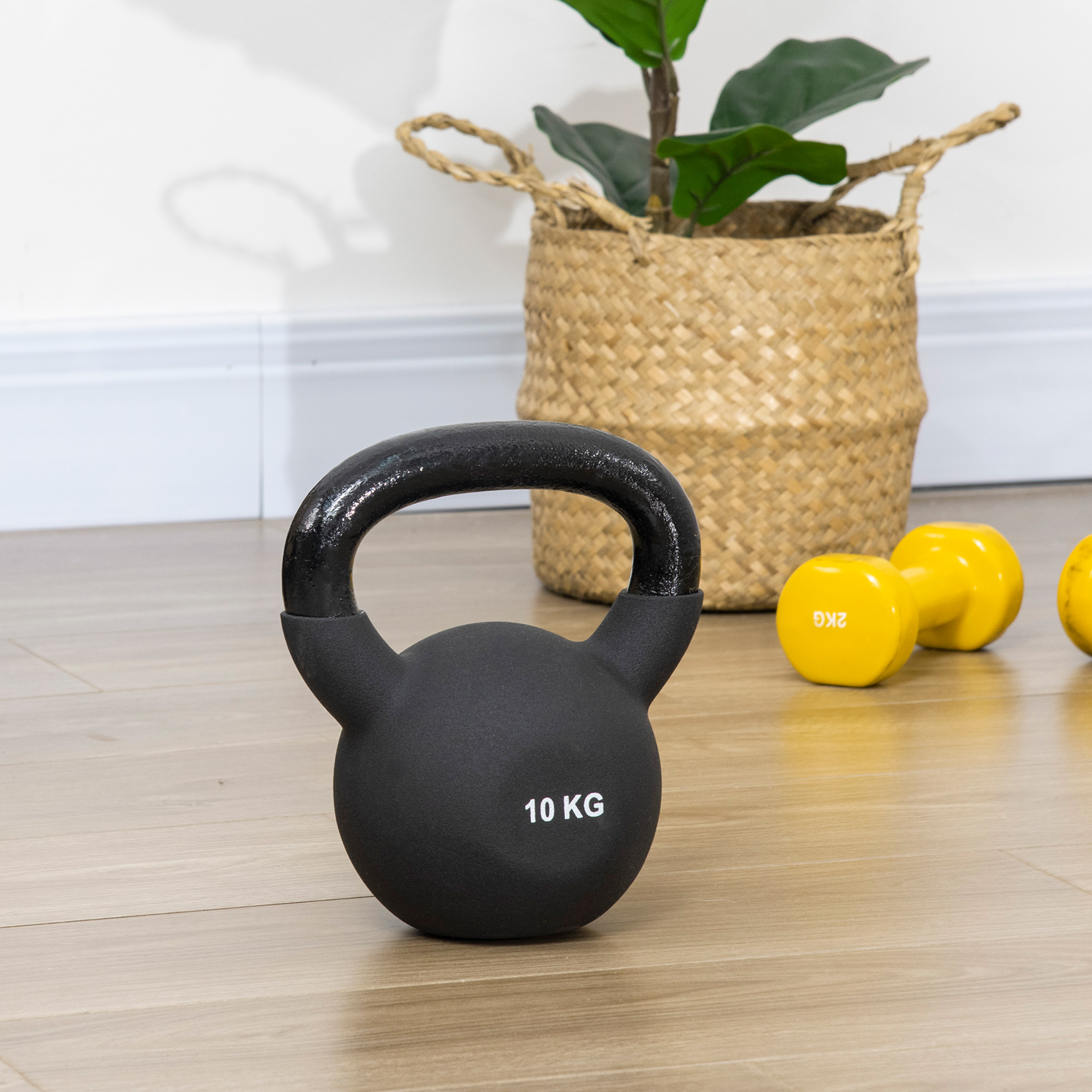 Eine schwarze Kettlebell mit der Aufschrift '10 KG', mit Kurzhanteln und einer Pflanze im Hintergrund.