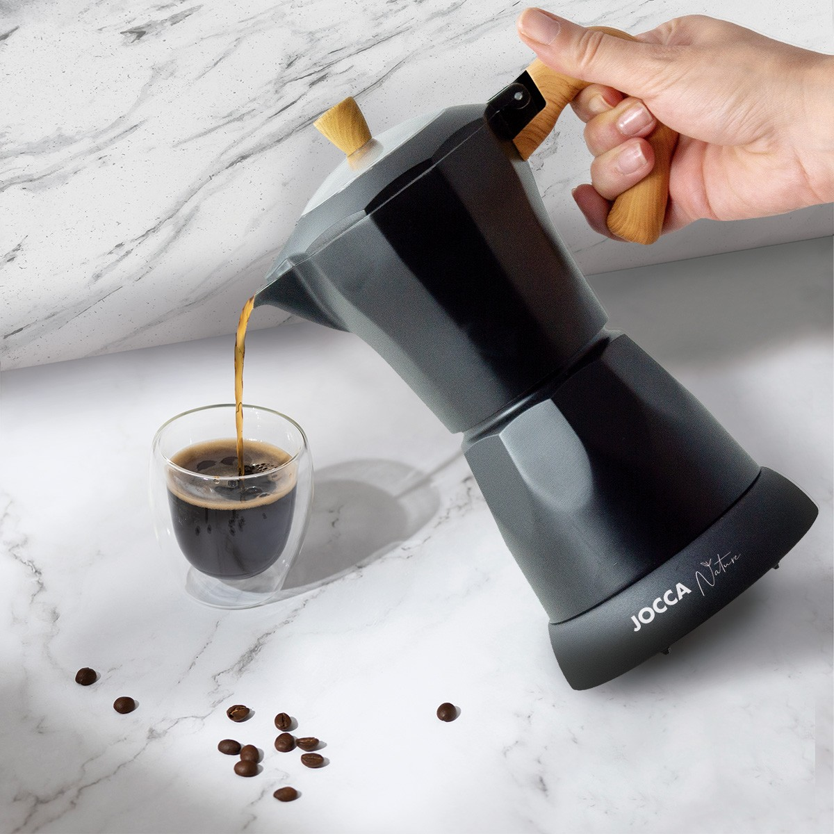 Eine Person gießt Kaffee aus einer dunkelgrauen Moka-Kanne in eine Glastasse, mit Kaffeebohnen auf der Oberfläche.