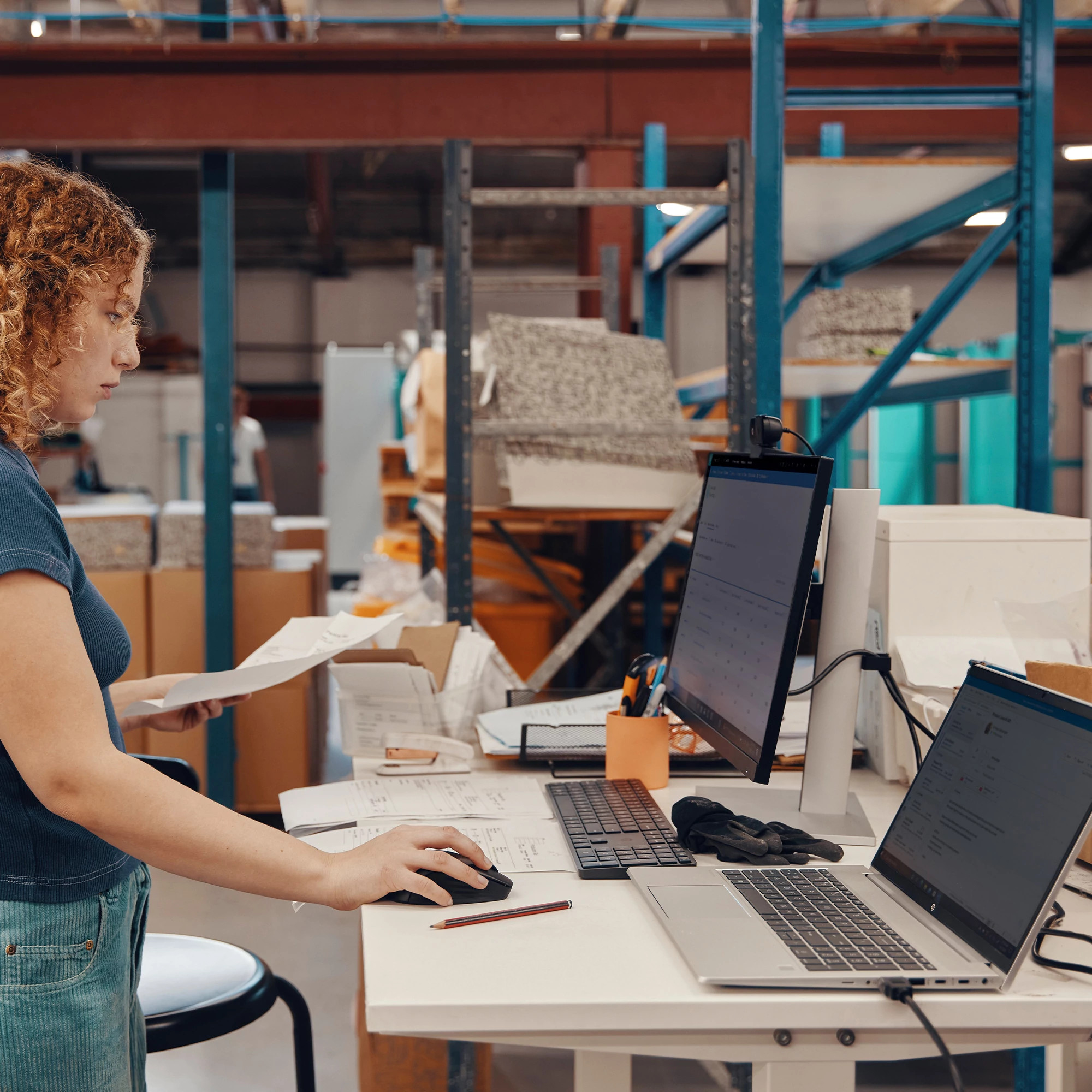 Kobieta w biurze magazynu korzystająca z laptopa i przeglądająca papiery.