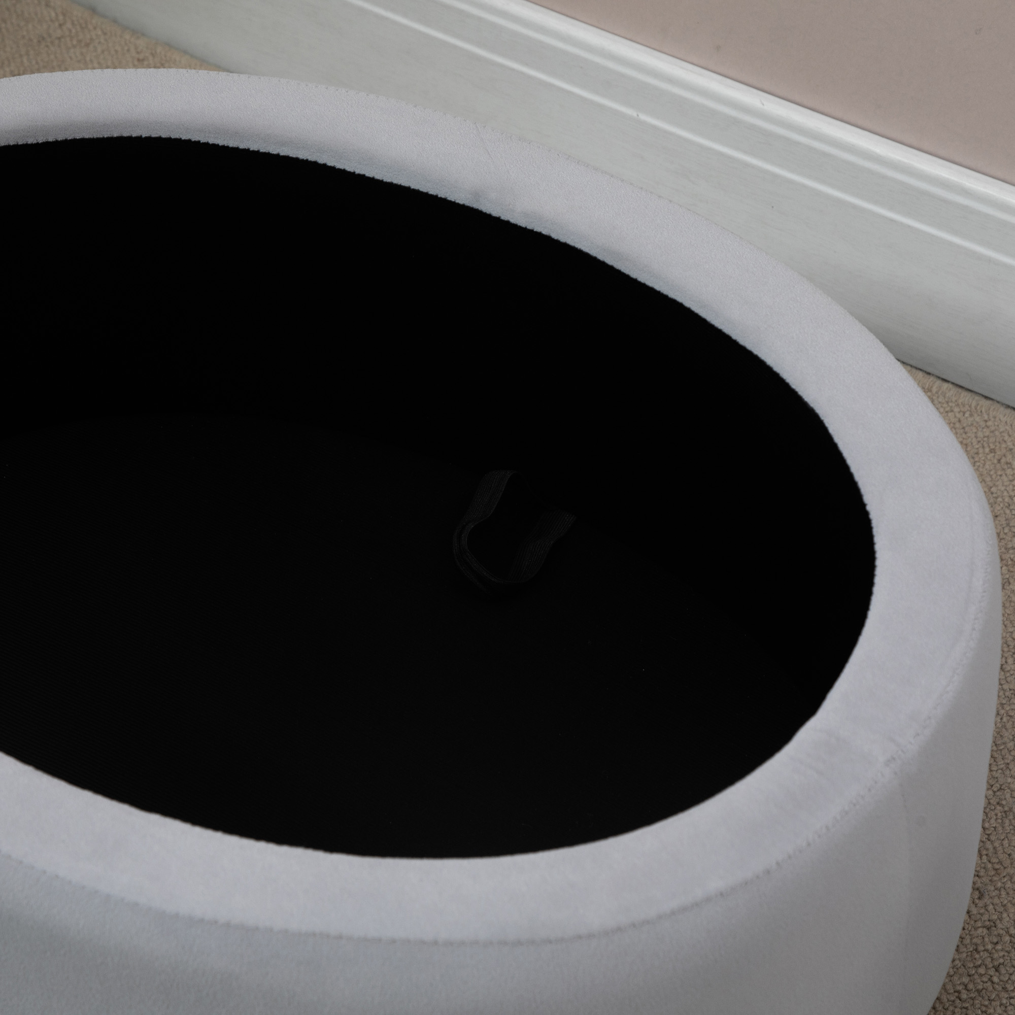 Weißer ovaler Hocker mit schwarzem Innenraum. Beige Teppich und weiße Wand sichtbar.